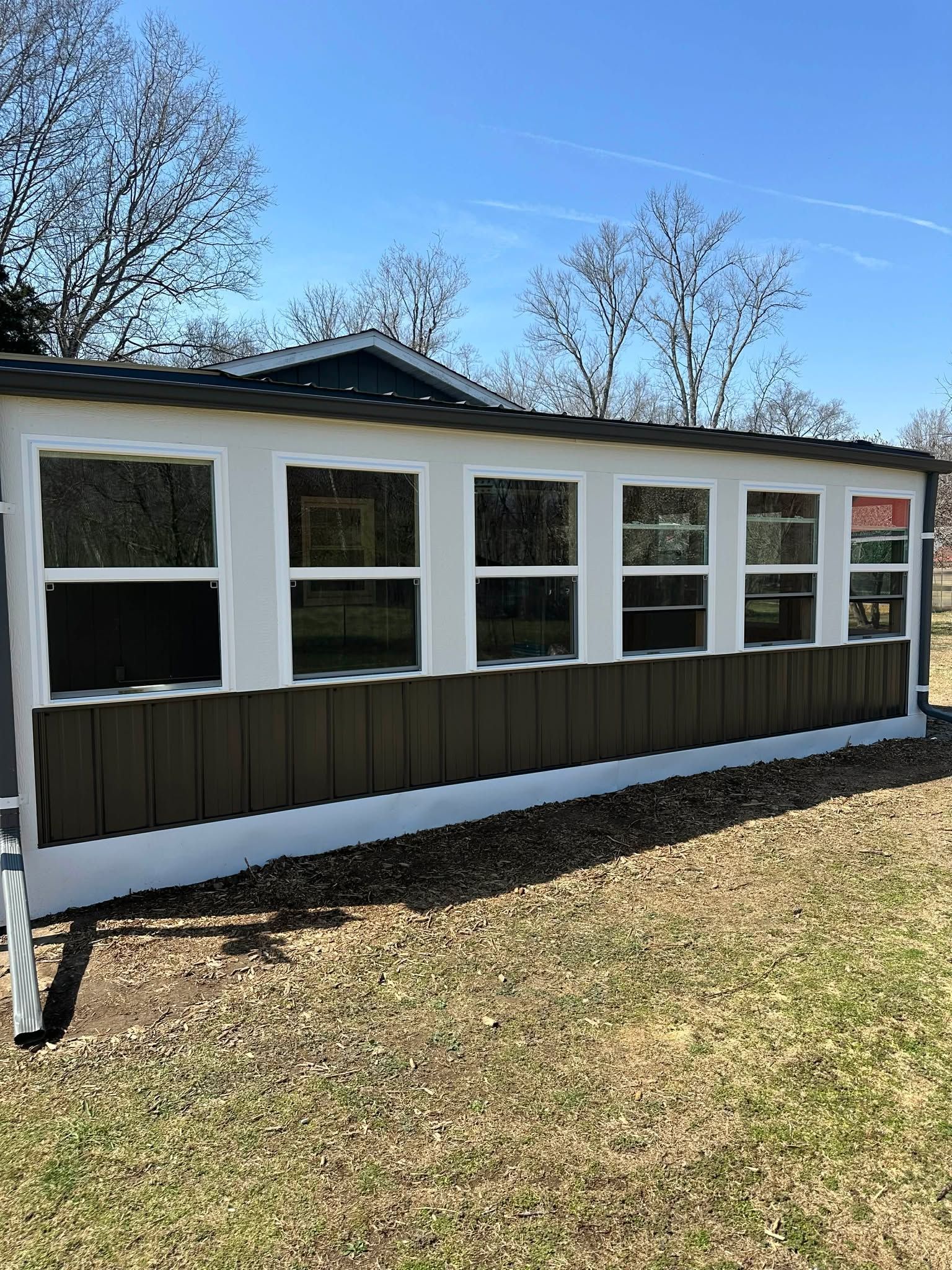 Small white building with multiple windows and dark siding, set on a grassy lot under a blue sky
