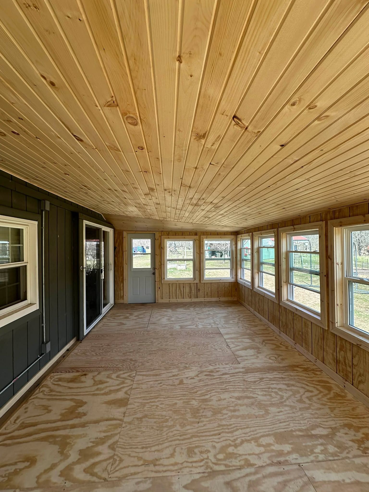 Unfinished enclosed porch with plywood floor, wood ceiling, and large windows along the right wall.