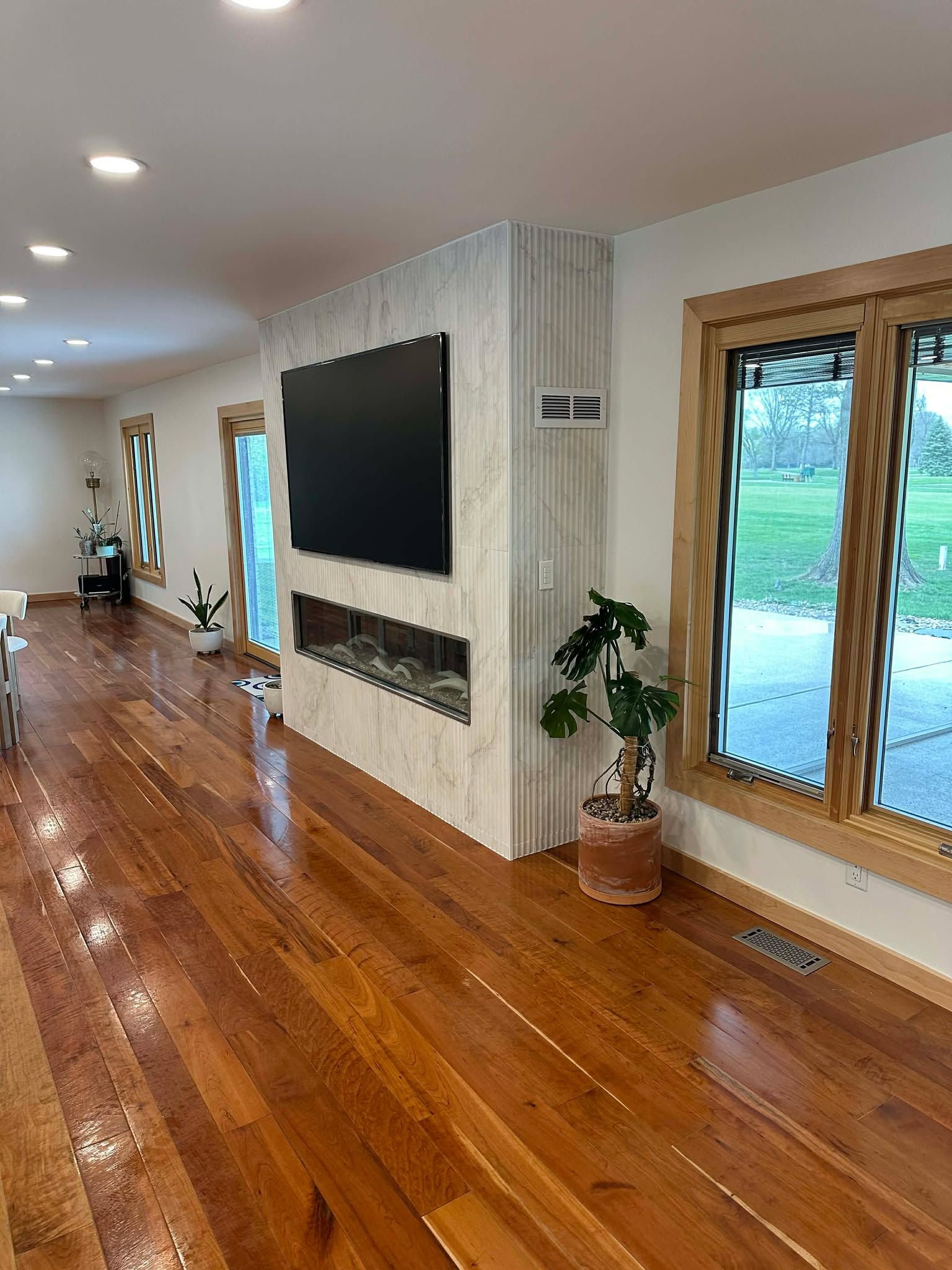 Empty living room with hardwood floors, wall-mounted TV, fireplace, and large windows overlooking greenery
