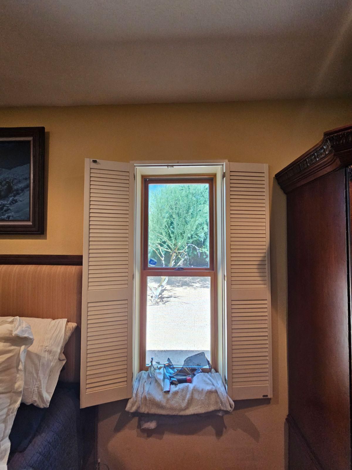 A window with open white plantation shutters in a bedroom, with a cloth covering the sill and furniture on either side.