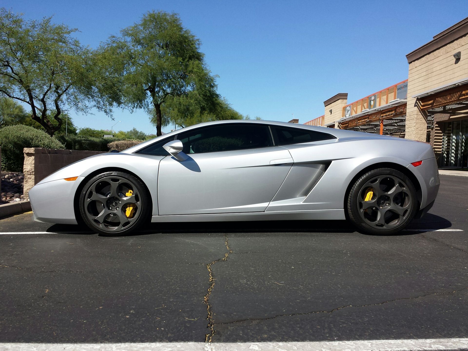 A silver Lamborghini Gallardo parked in an asphalt lot on a sunny day.