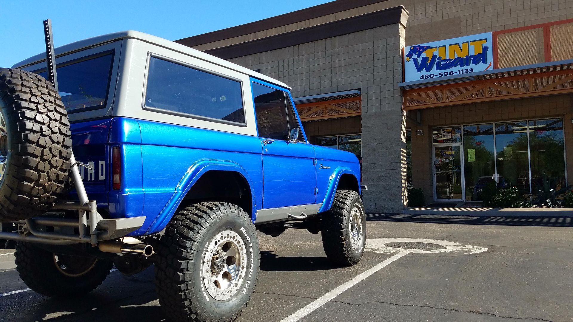 A bright blue Ford Bronco with a white roof and off-road tires parked in front of a Tint Wizard shop on a sunny day.