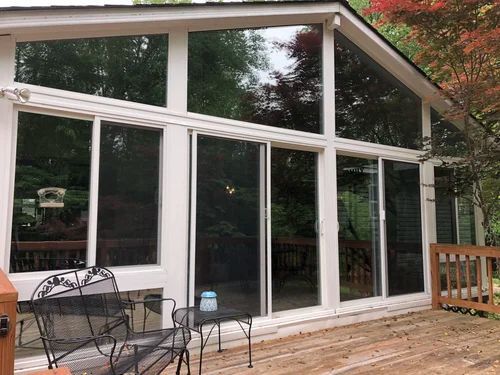Modern glass sunroom with white trim, deck chairs, and a wooden deck overlooking trees