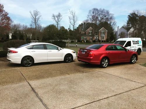 Two parked cars, white sedan and red sedan, on a driveway with houses and trees in the background