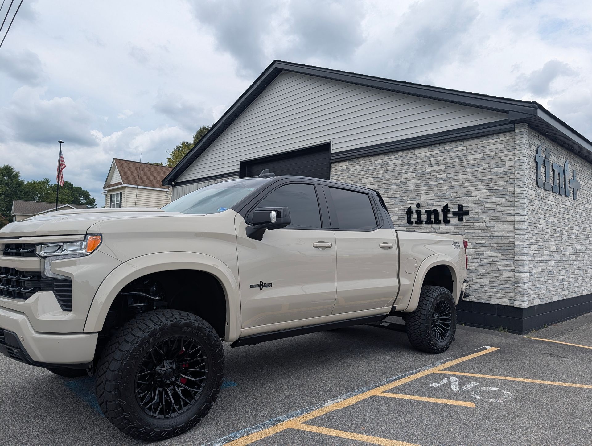 White lifted pickup truck parked outside a white building with “tint+” signage on a cloudy day