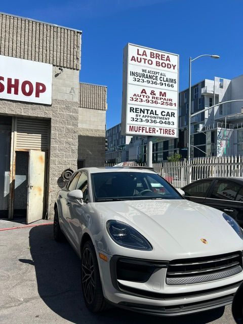 White Porsche SUV parked outside an auto body shop with a sign for rentals, mufflers, and tires.