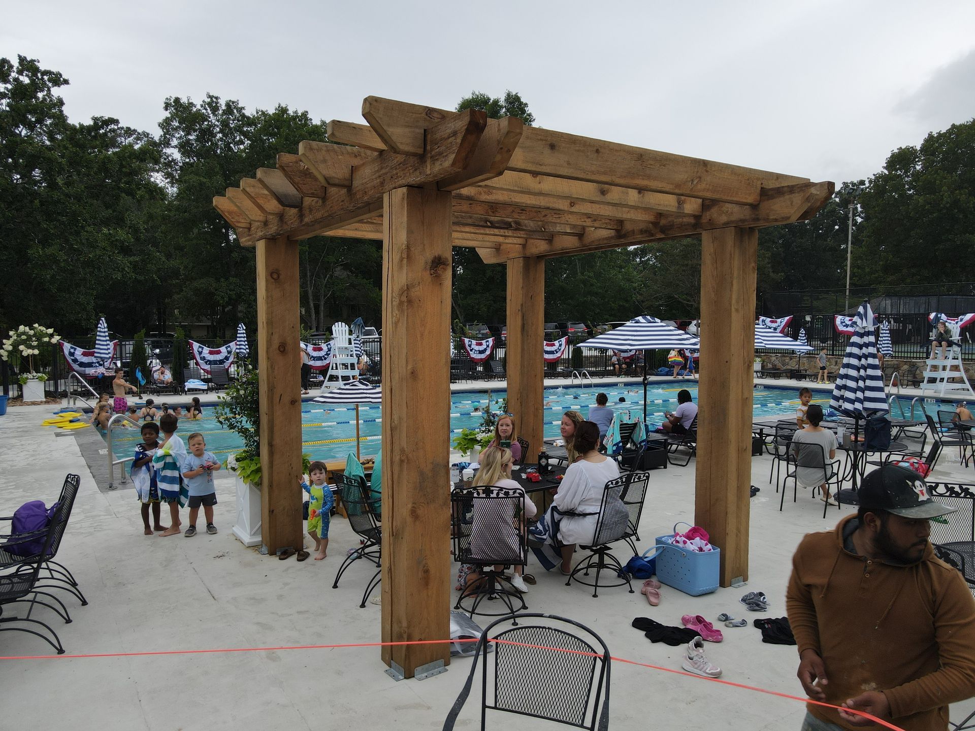 A group of people are sitting under a wooden pergola near a swimming pool.