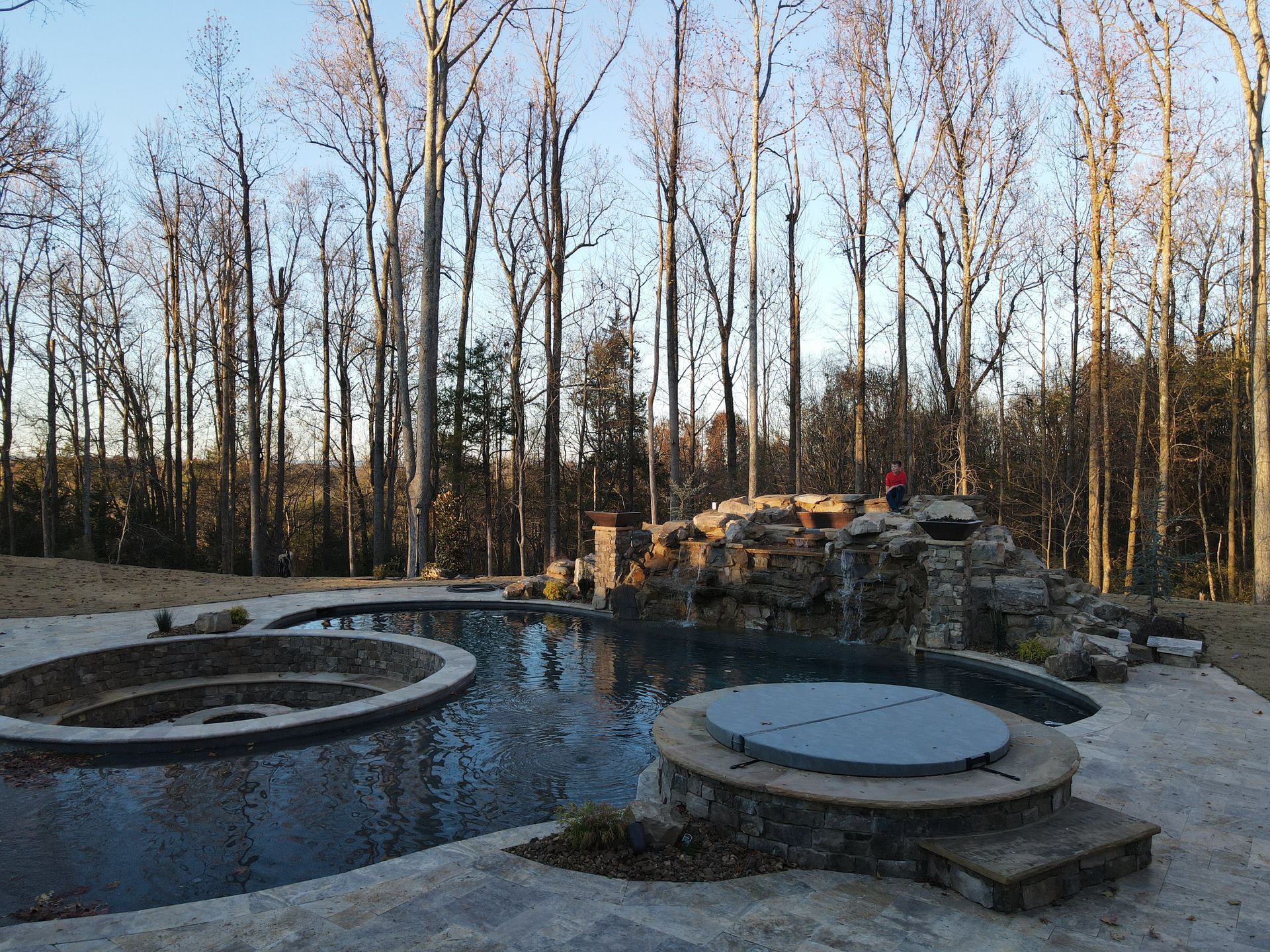 A large swimming pool with a hot tub in the middle of it surrounded by trees.