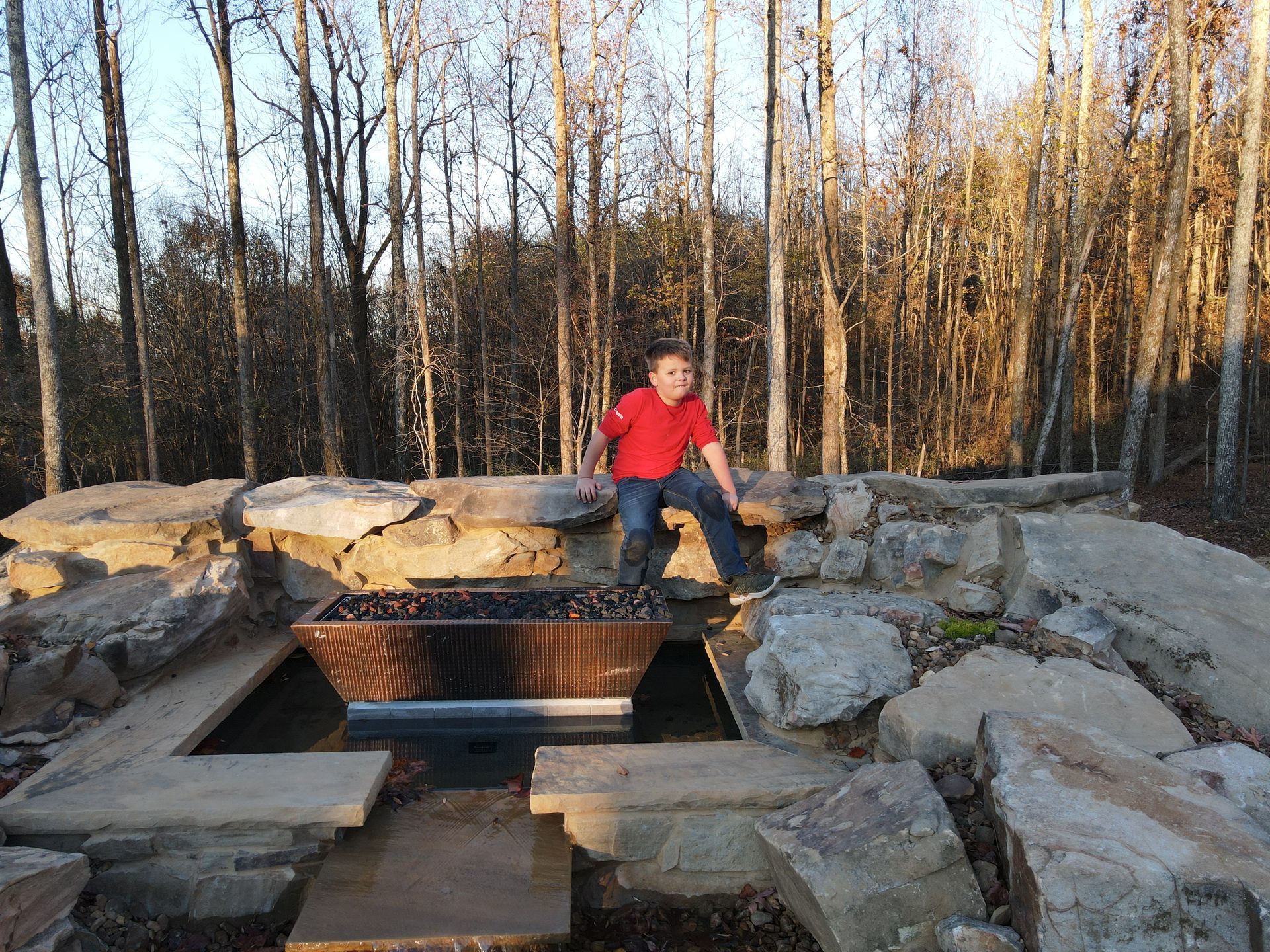 A young boy is sitting on a rock near a fire pit in the woods.