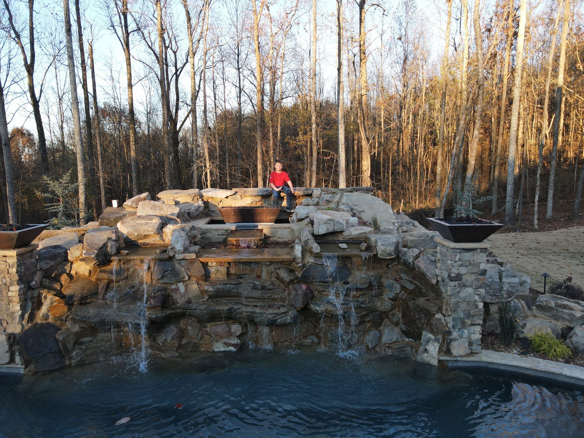 A man in a red shirt is standing on top of a waterfall next to a pool.