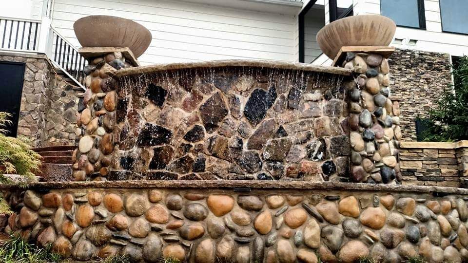 A large stone wall with a waterfall in the middle of it in front of a house.