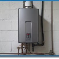 Gray tankless water heater mounted on a cinder block wall with visible plumbing pipes