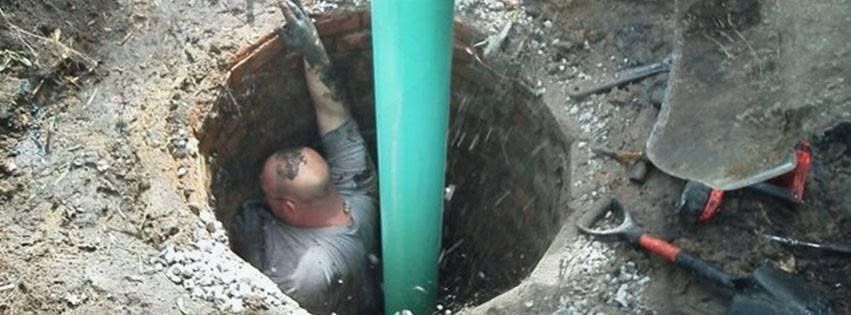 Worker in a trench installing a turquoise pipe beside dirt, with tools on the ground.