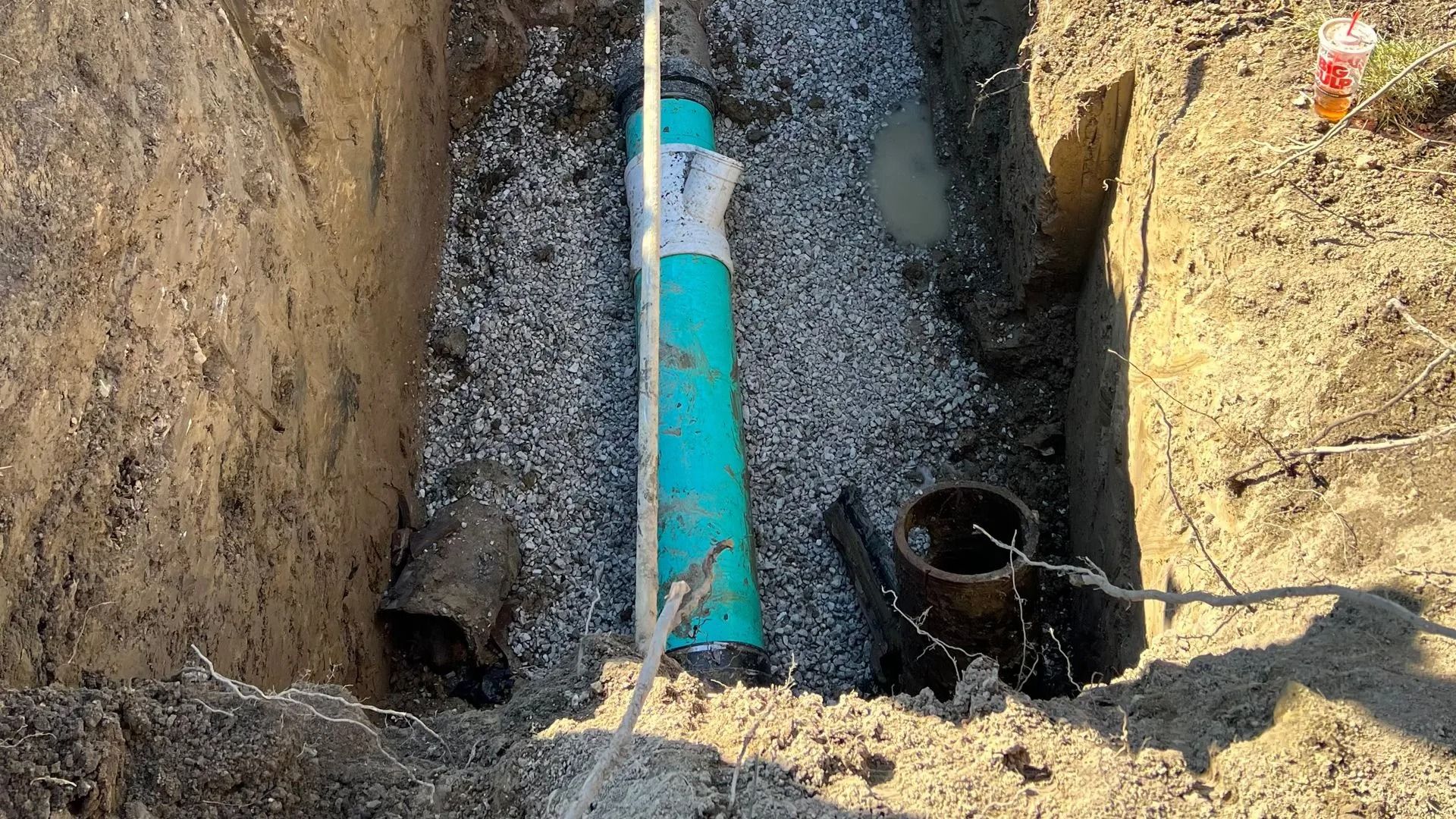 Trench with a turquoise pipe and black bucket in gravel between dirt walls