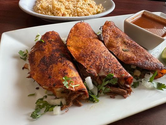 Three crispy tacos with meat and cilantro, rice, and dipping sauce on a white plate.
