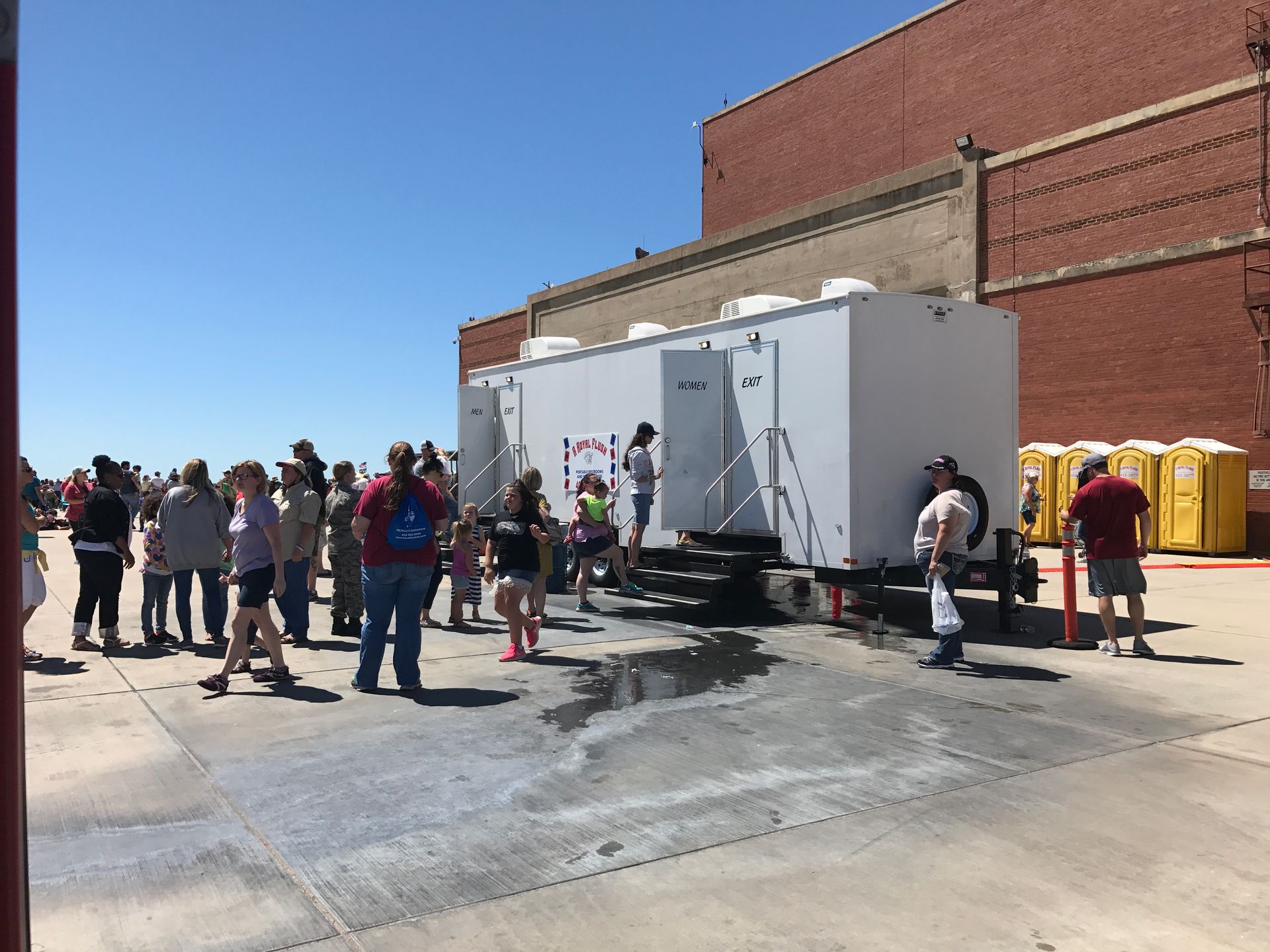 People gather near a white portable restroom trailer and yellow portable toilets on a sunny day.