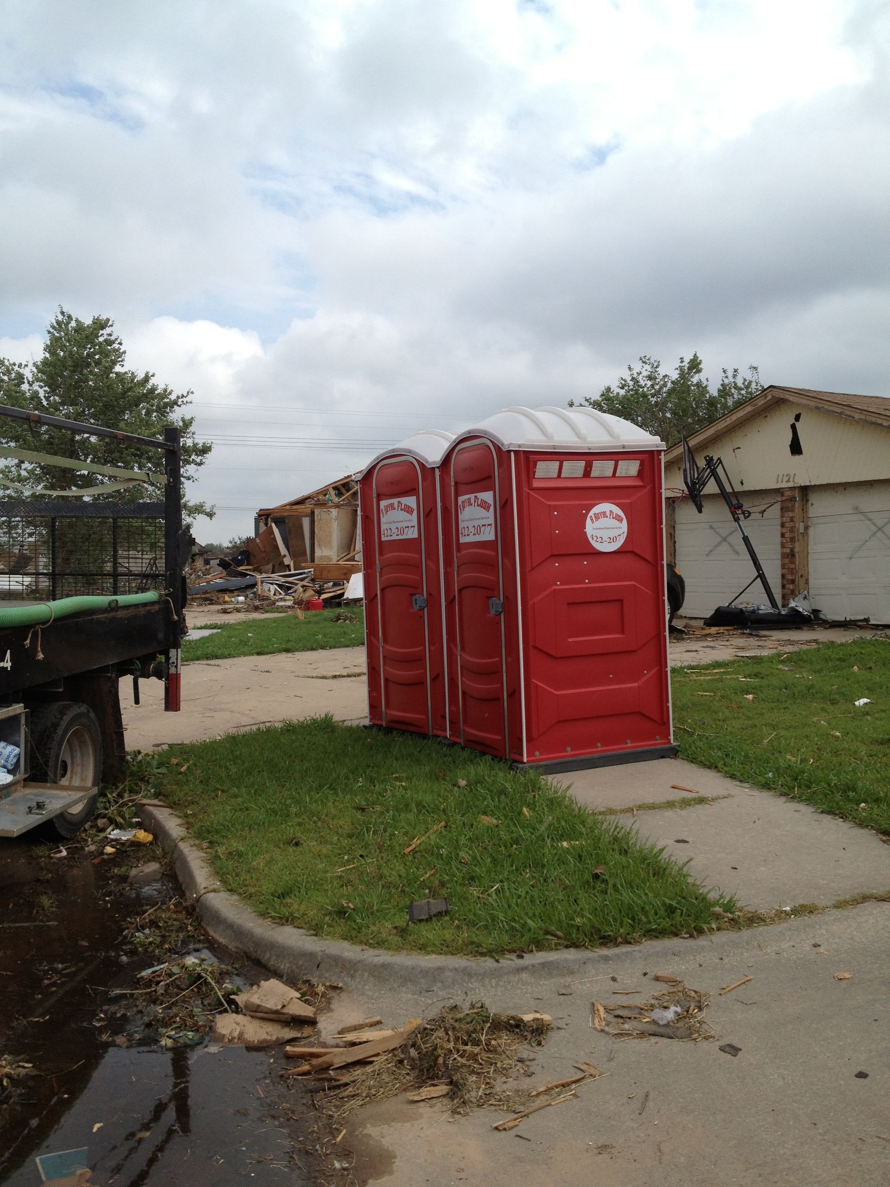 Two red portable toilets on a sidewalk in front of damaged buildings; debris scattered around.
