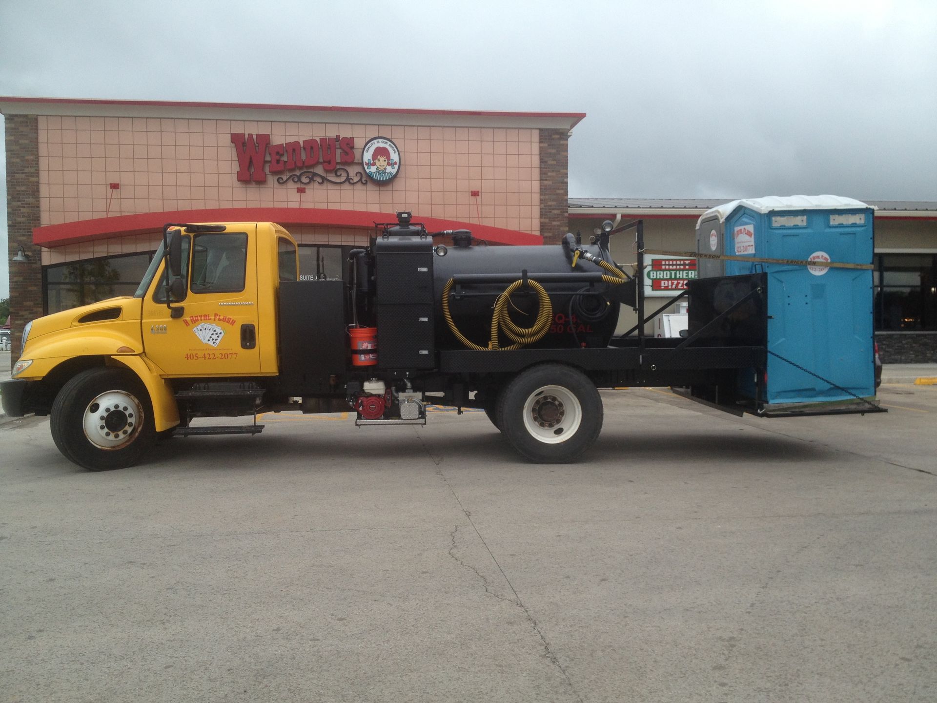 Yellow sanitation truck parked in front of a Waynes BBQ restaurant, carrying a blue portable toilet.