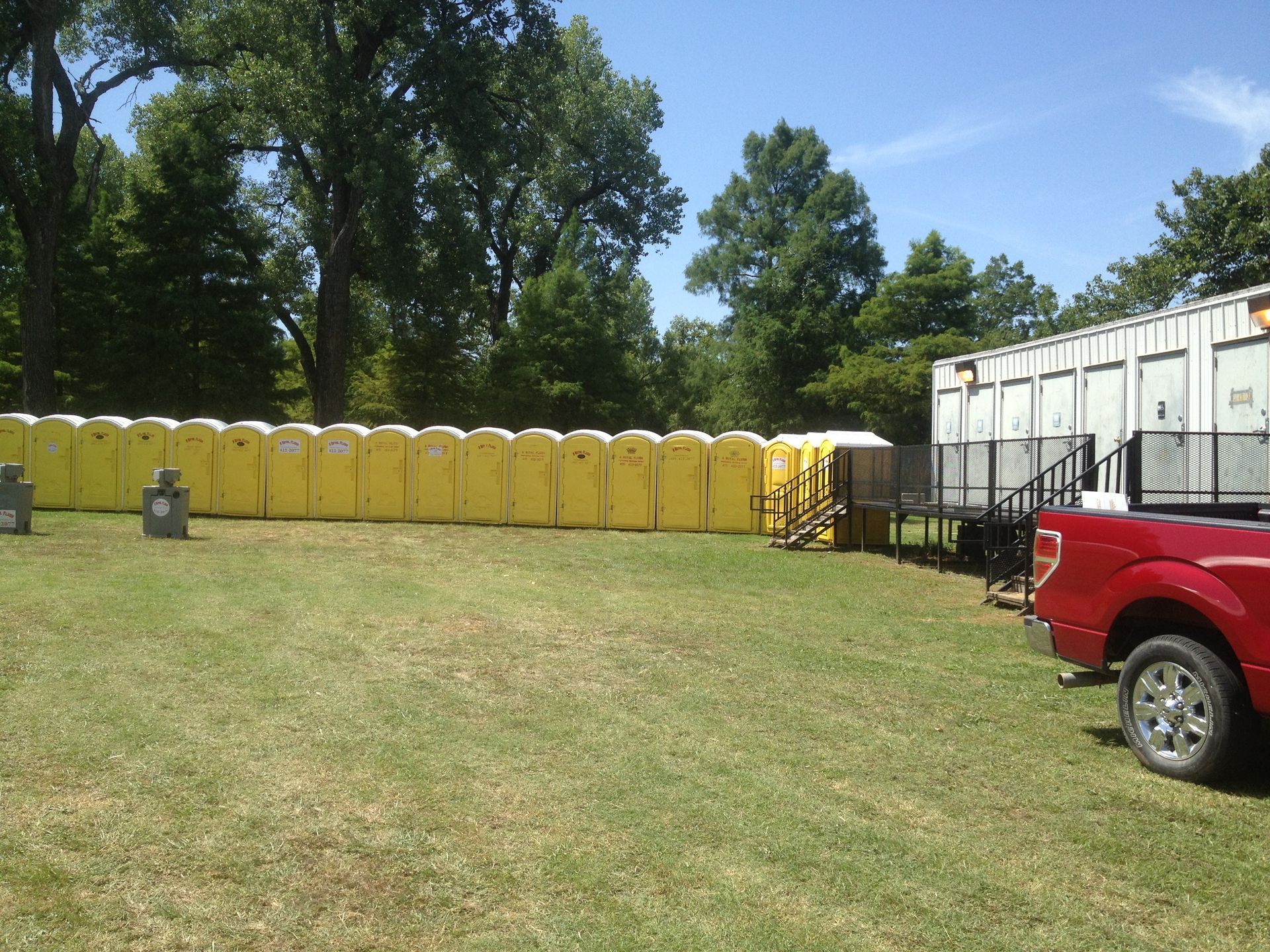 Row of yellow portable toilets in a grassy area, alongside a white trailer and a red truck.