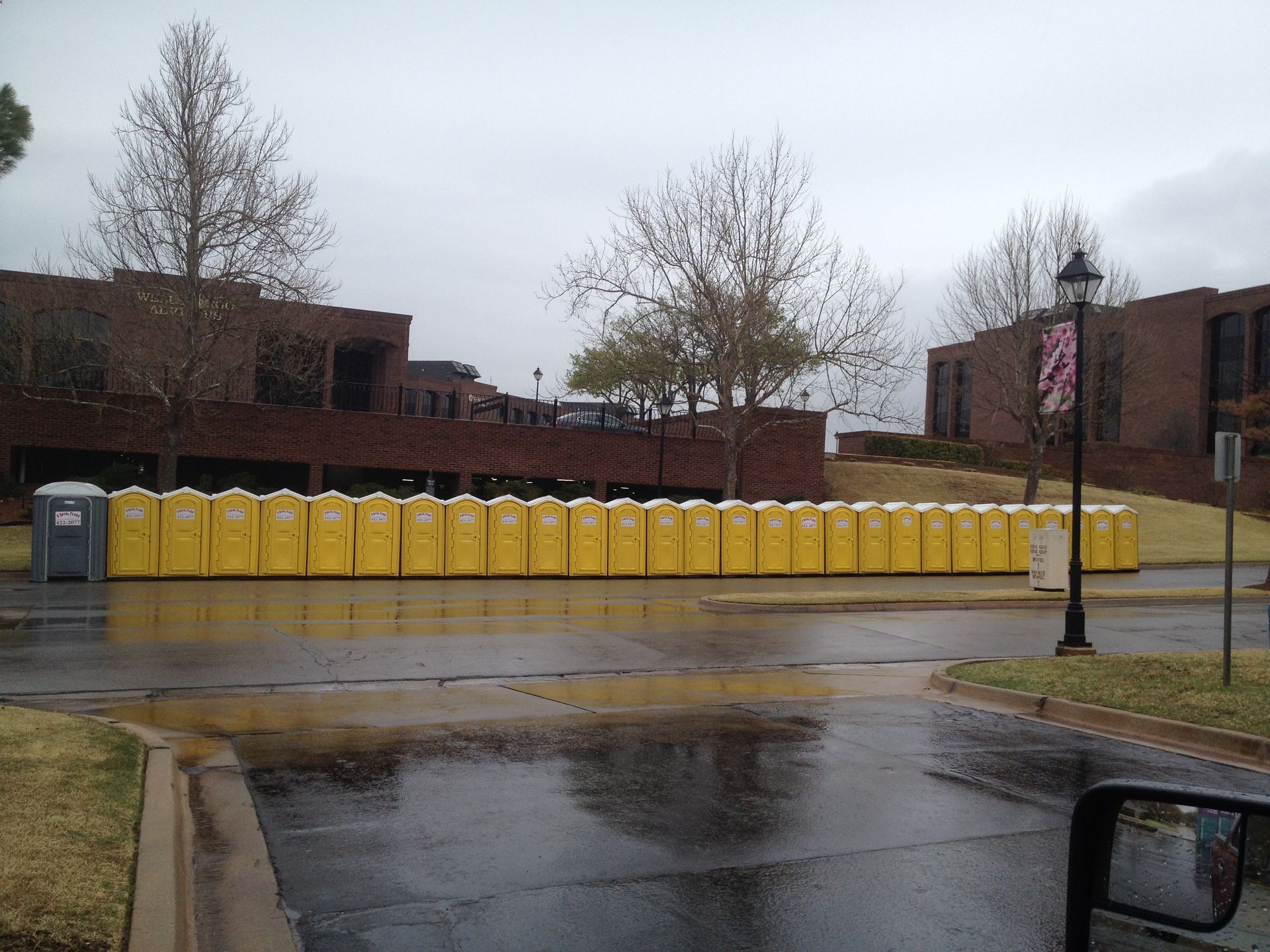 A long row of yellow portable toilets in front of a brick building on a wet, overcast day.