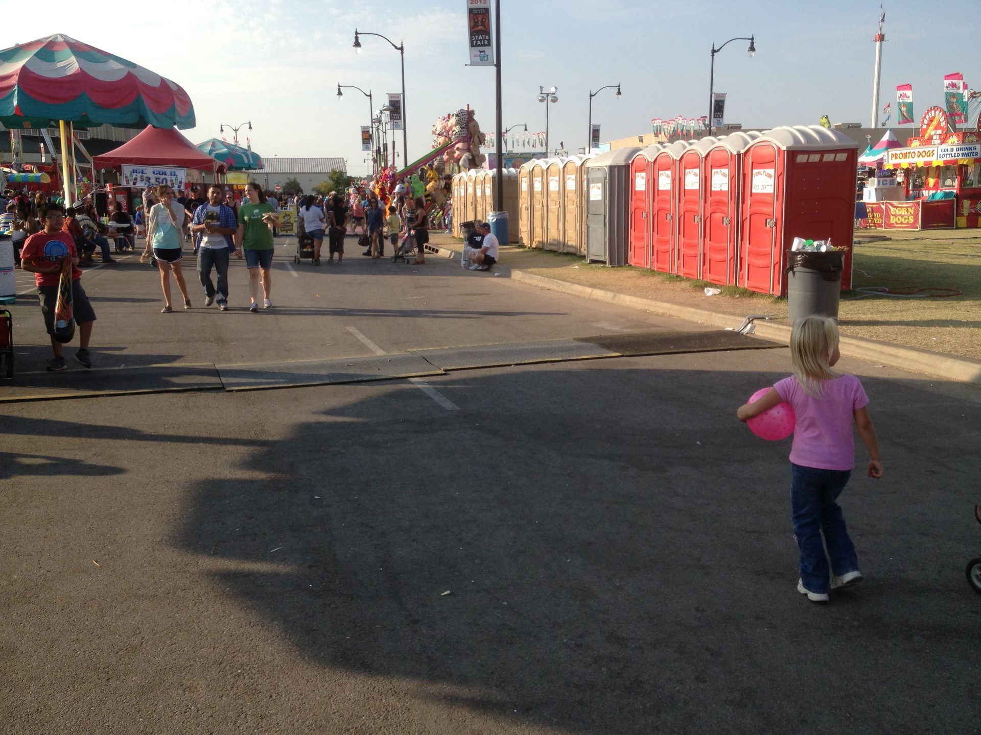 Girl with a pink ball walks on a paved road at a fair, with port-a-potties and crowds in the background.