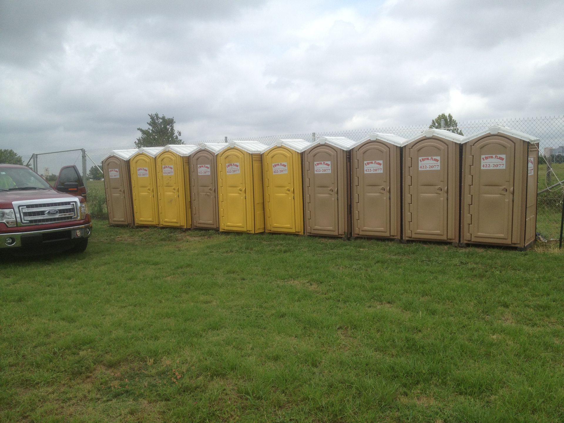 A row of portable toilets on green grass next to a red truck under a cloudy sky.