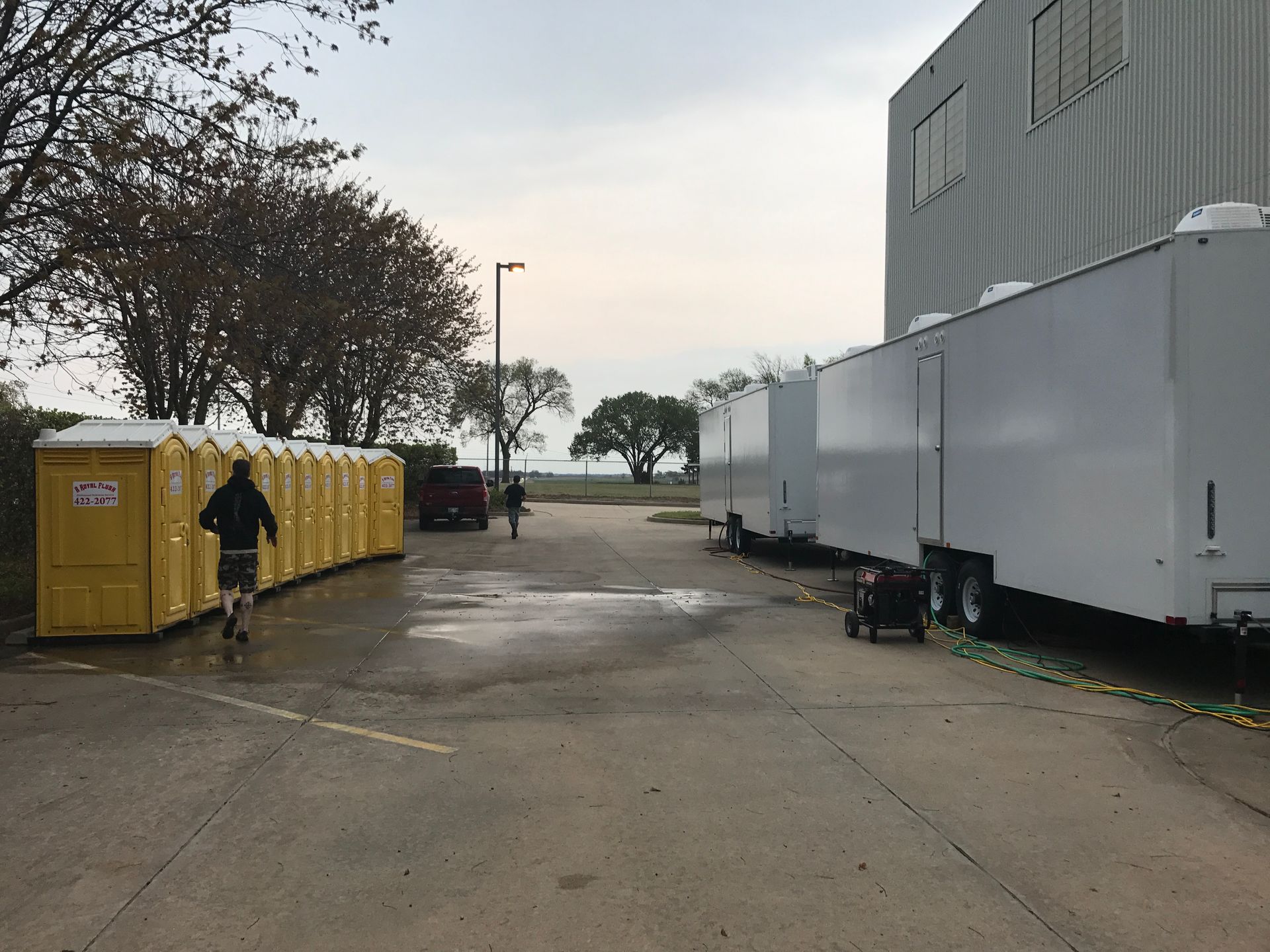 Port-a-potties and trailers lined up outdoors. Person runs toward them, another walks away. Cloudy day.