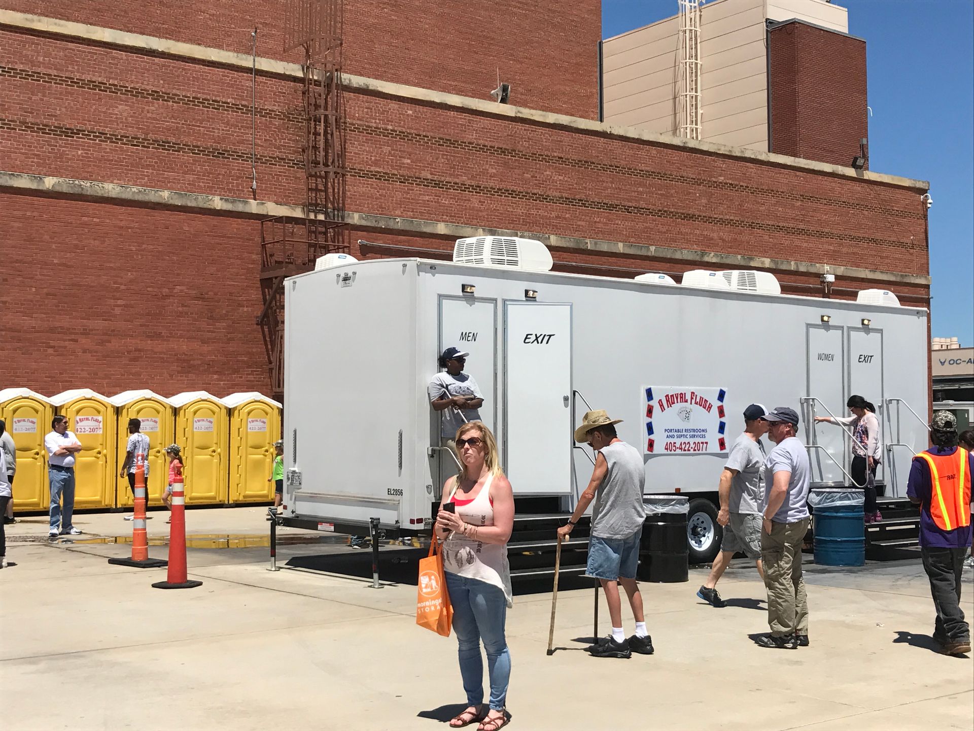 People line up near a trailer with restrooms next to a brick building. Several portable toilets in yellow are also visible.