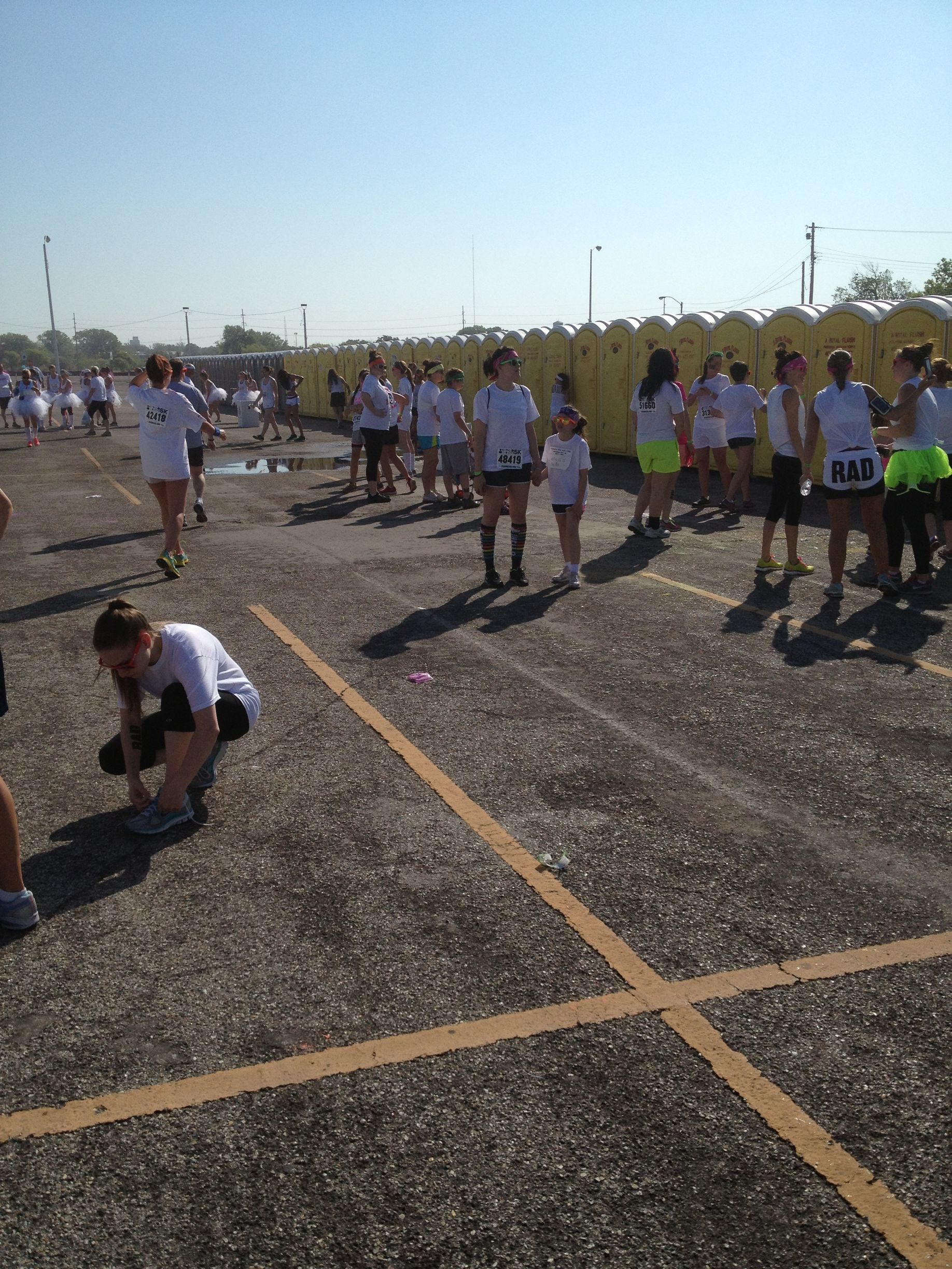 People in white shirts at an outdoor event near portable toilets; some are running, one is tying a shoe.