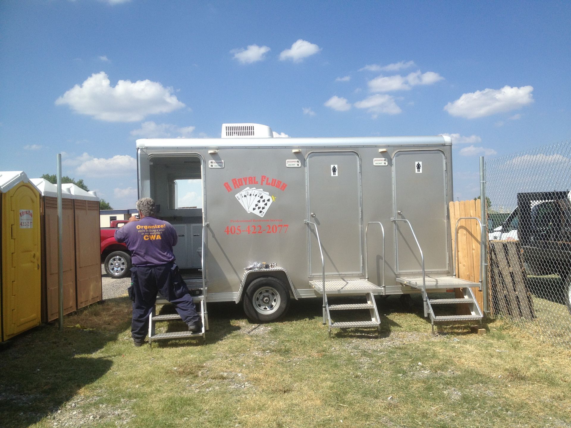 Portable restroom trailer with steps, worker, and portable toilets on a grassy field.