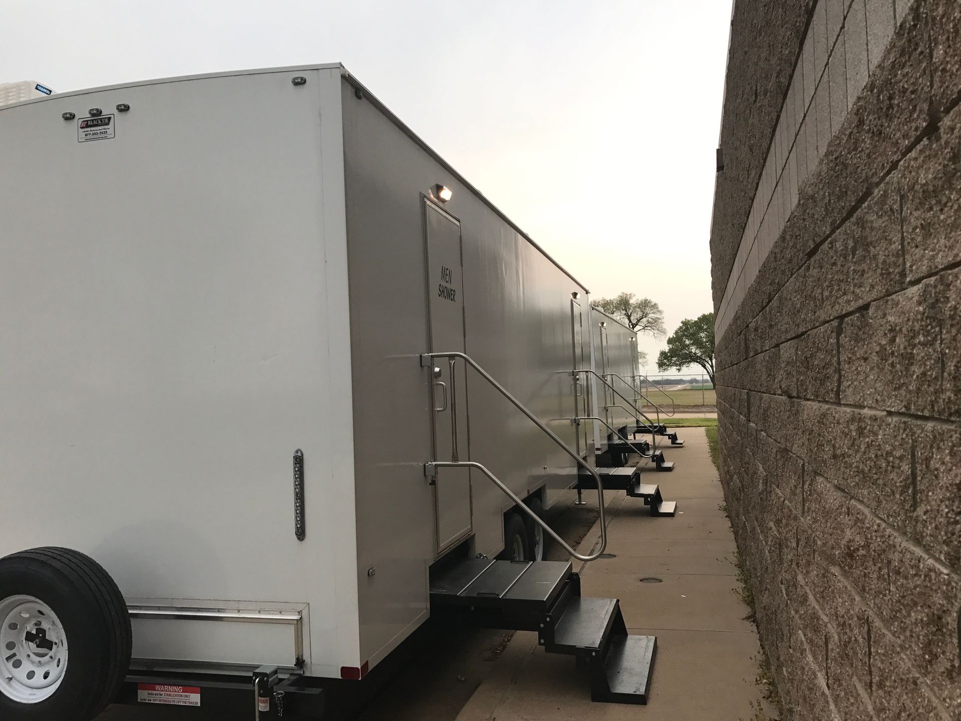 White portable restroom trailers lined up next to a brick wall on a concrete surface.