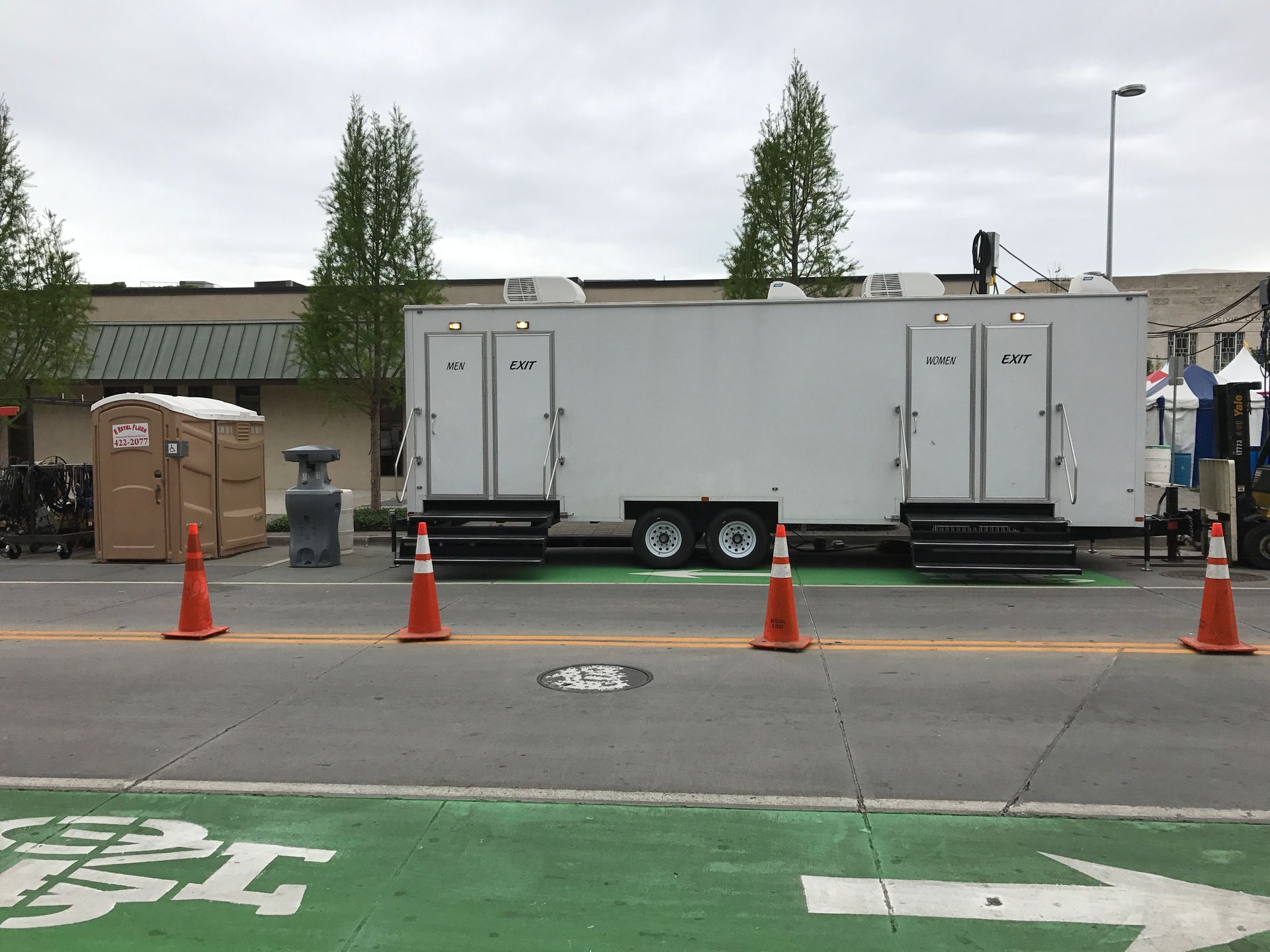 Mobile restrooms on a city street, orange cones in front, with trees and a building in the background.
