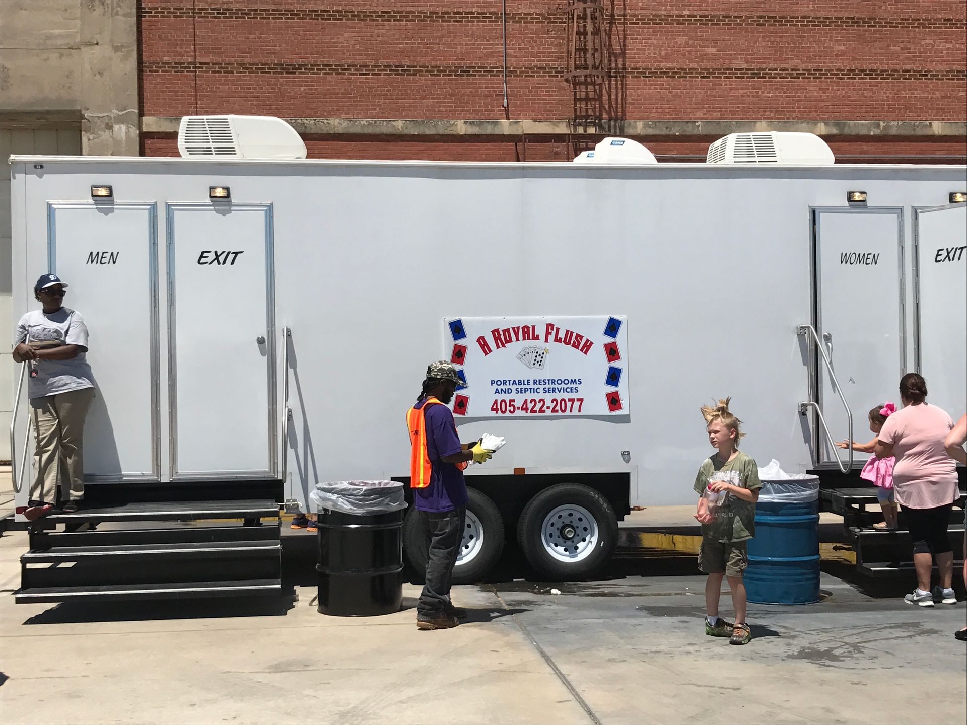Portable restroom trailer with people in front of it. A person stands on the steps, others nearby.