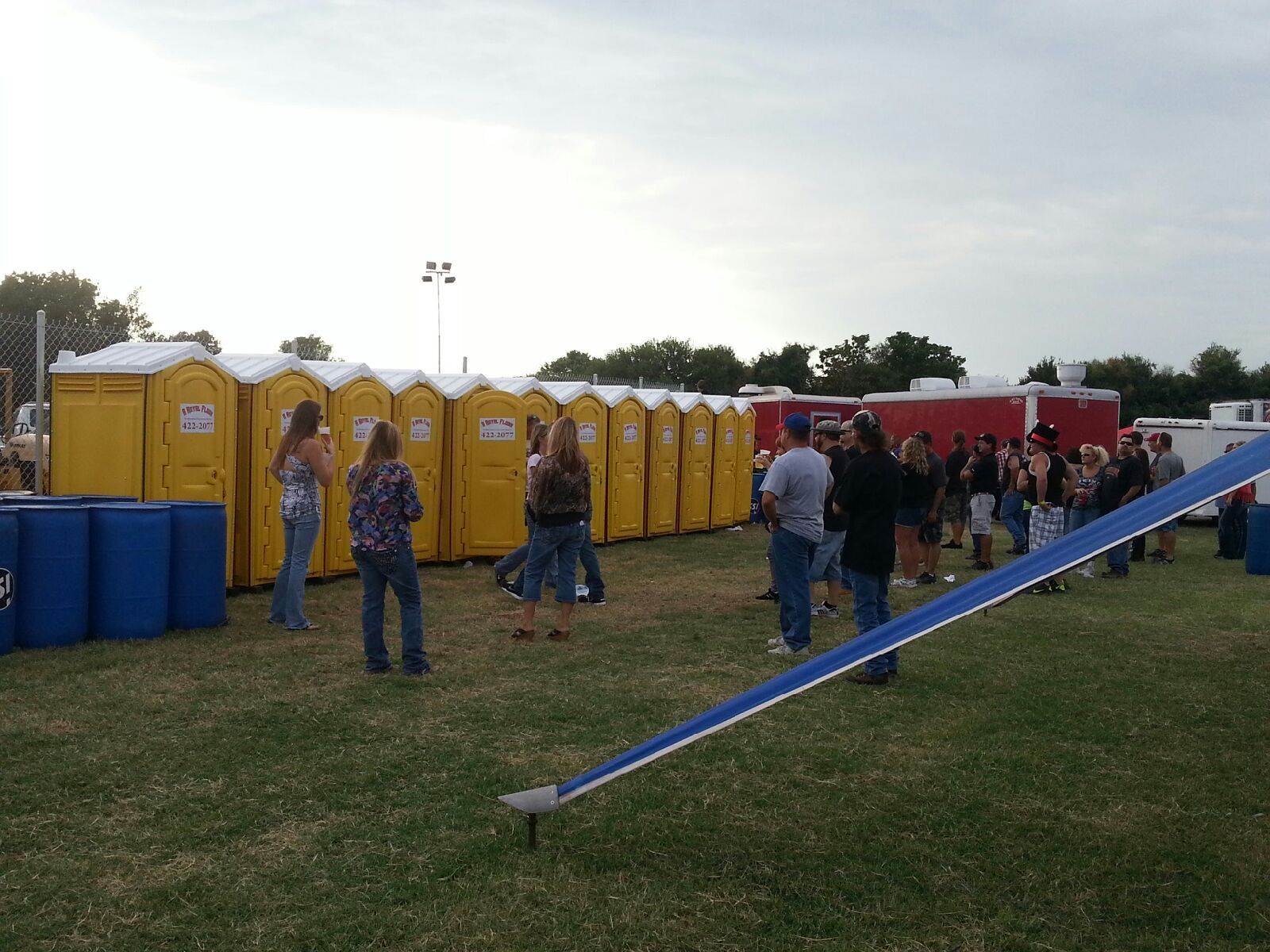 People waiting in line for yellow portable toilets in a grassy area.