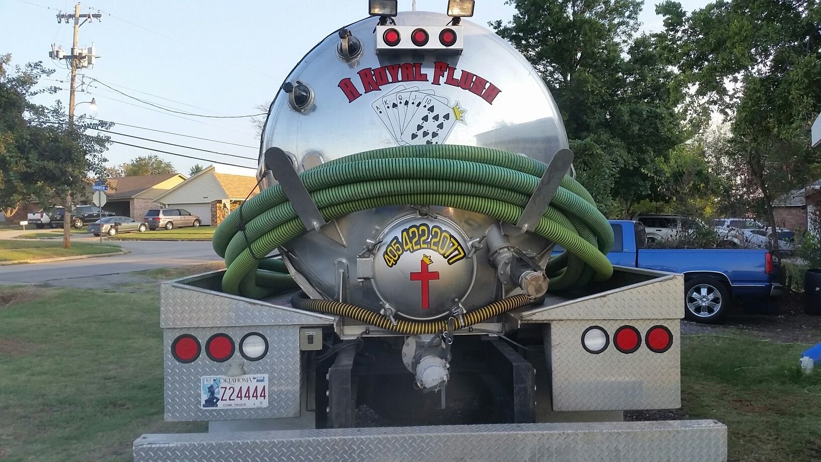 Rear view of a silver septic tank truck. Green hose wraps around a cross logo. Red taillights are visible.