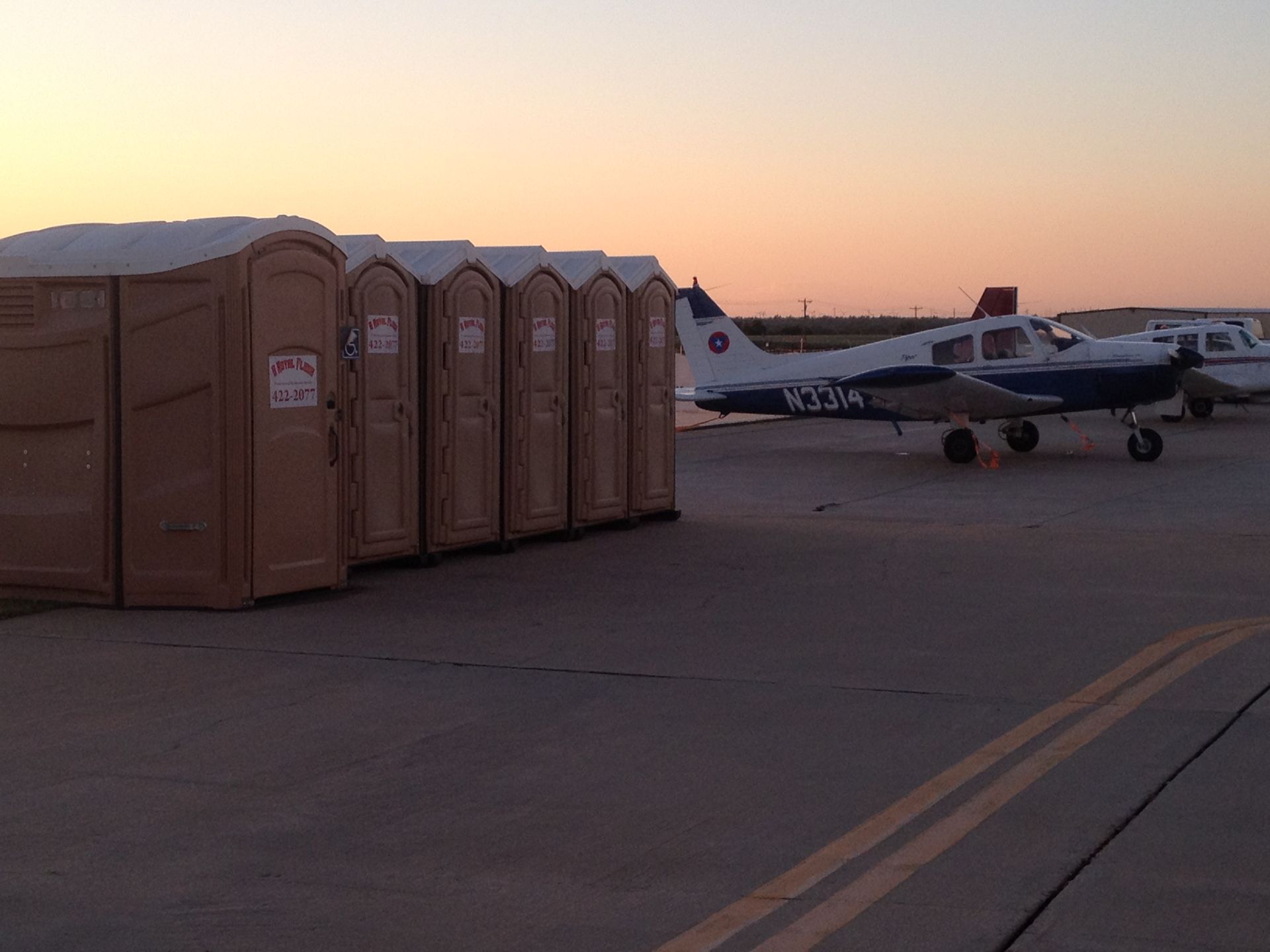 Row of portable toilets near a small airplane on a tarmac at sunset.