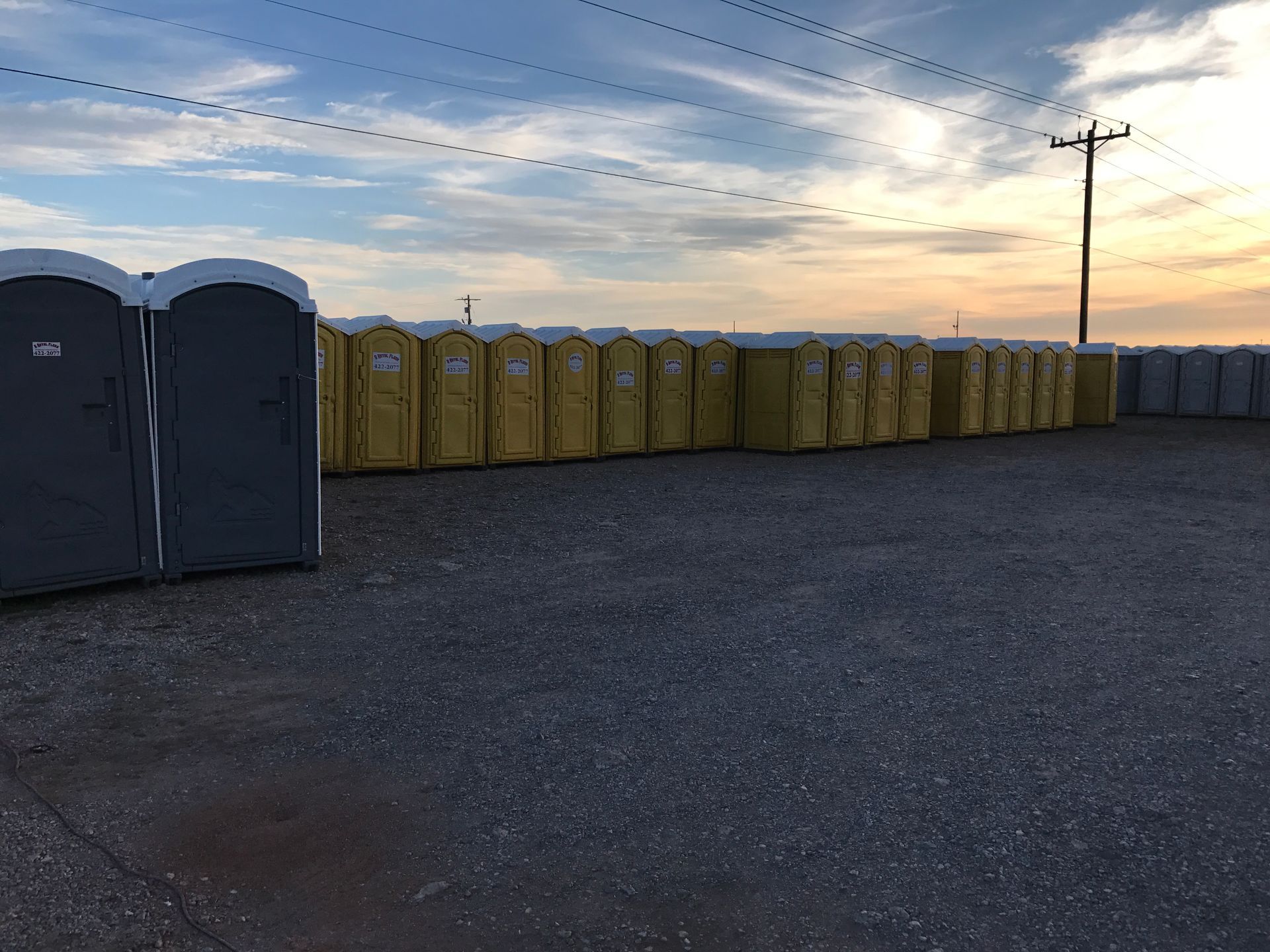 Rows of yellow and gray portable toilets in a gravel lot under a cloudy sky.