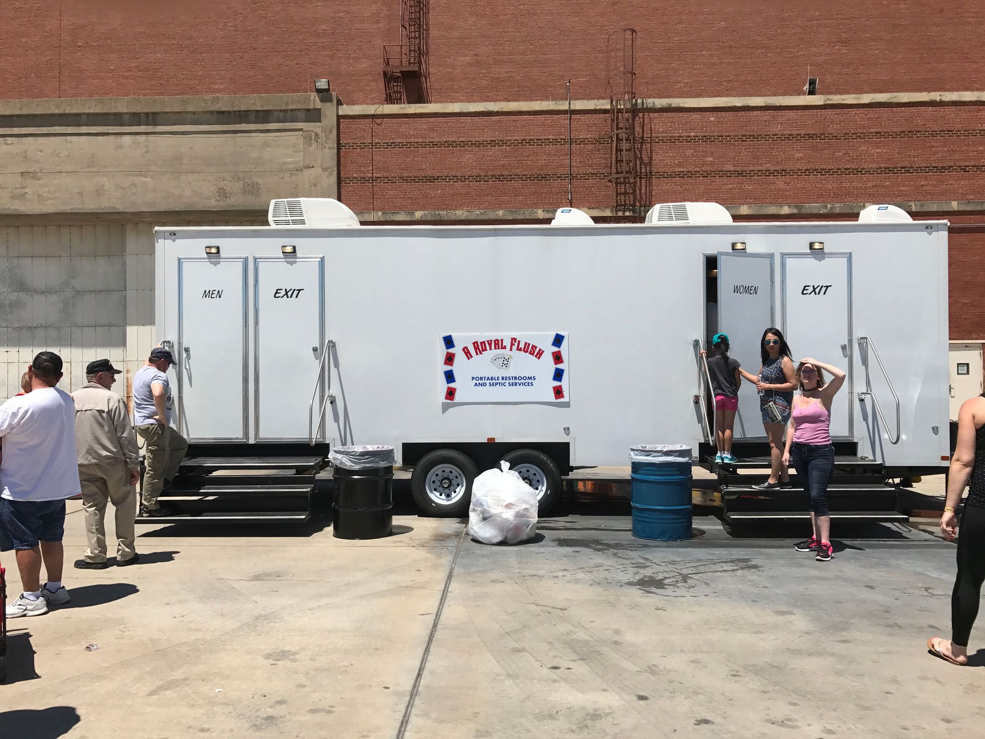 White portable restroom trailer with people in front of brick building. Some enter/exit the trailer.