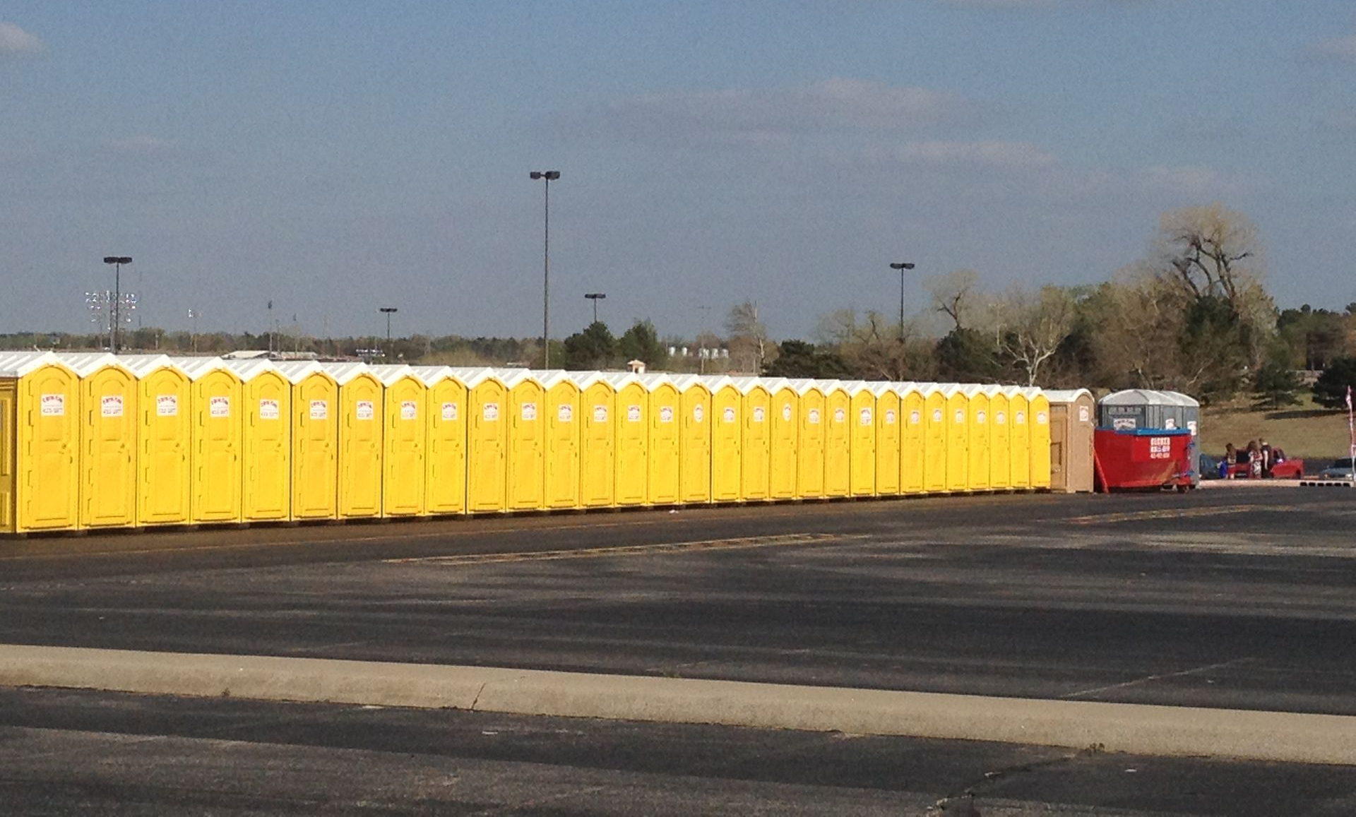 Long row of yellow portable toilets in a parking lot. Red and white trailer at the end. Blue sky.