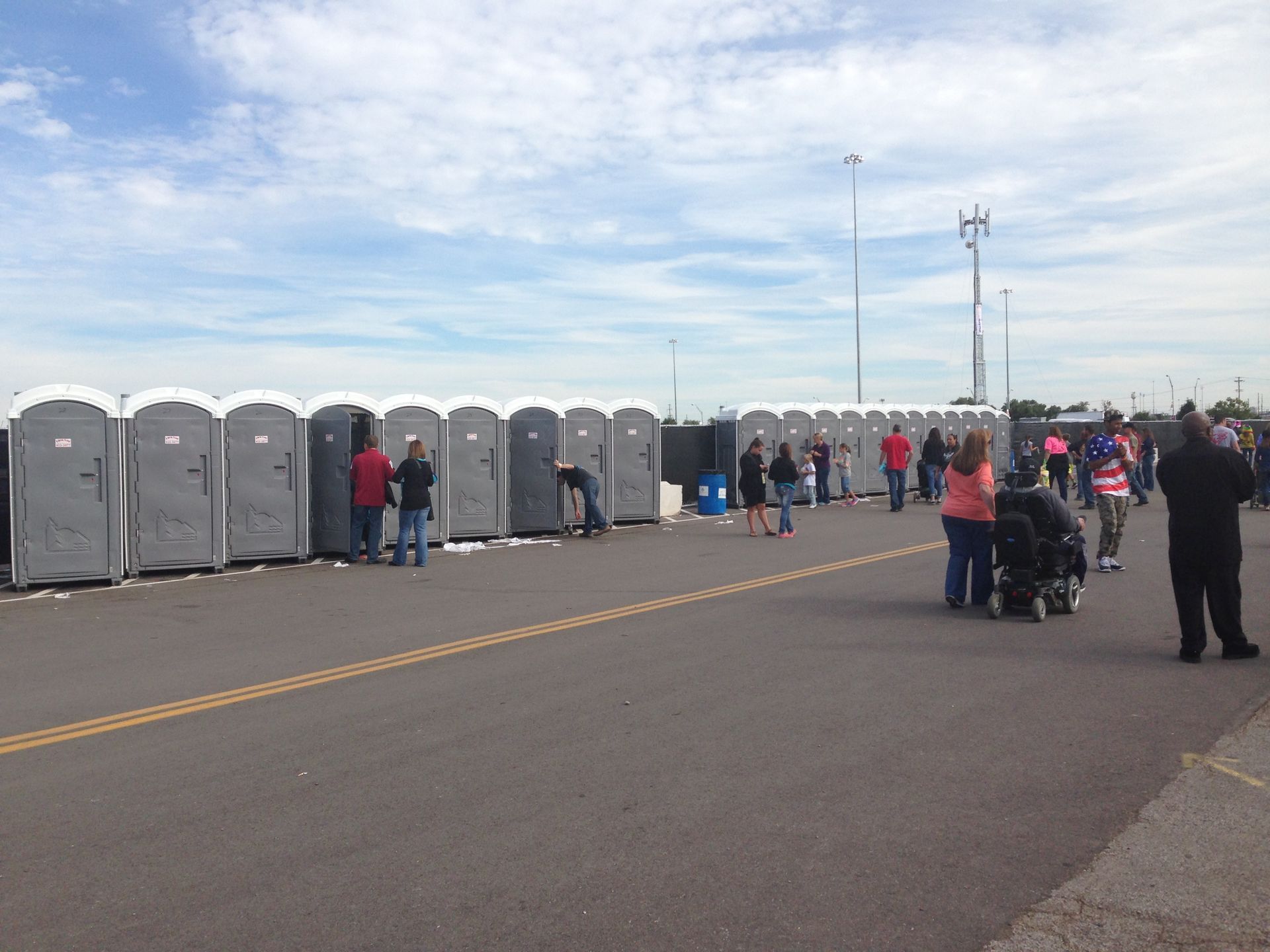 Row of portable toilets with people in line on an asphalt surface under a cloudy sky.