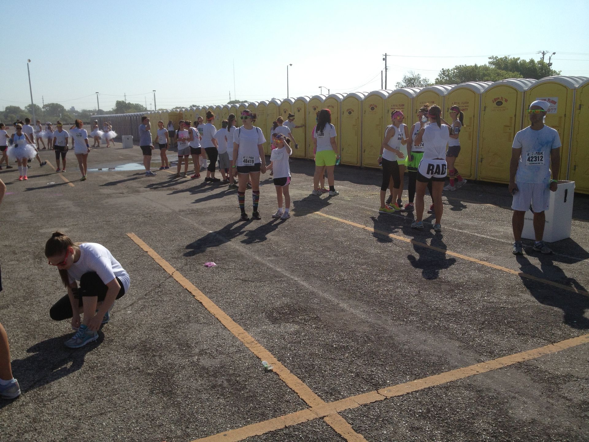 People in white shirts and shorts at a race, standing near portable toilets.