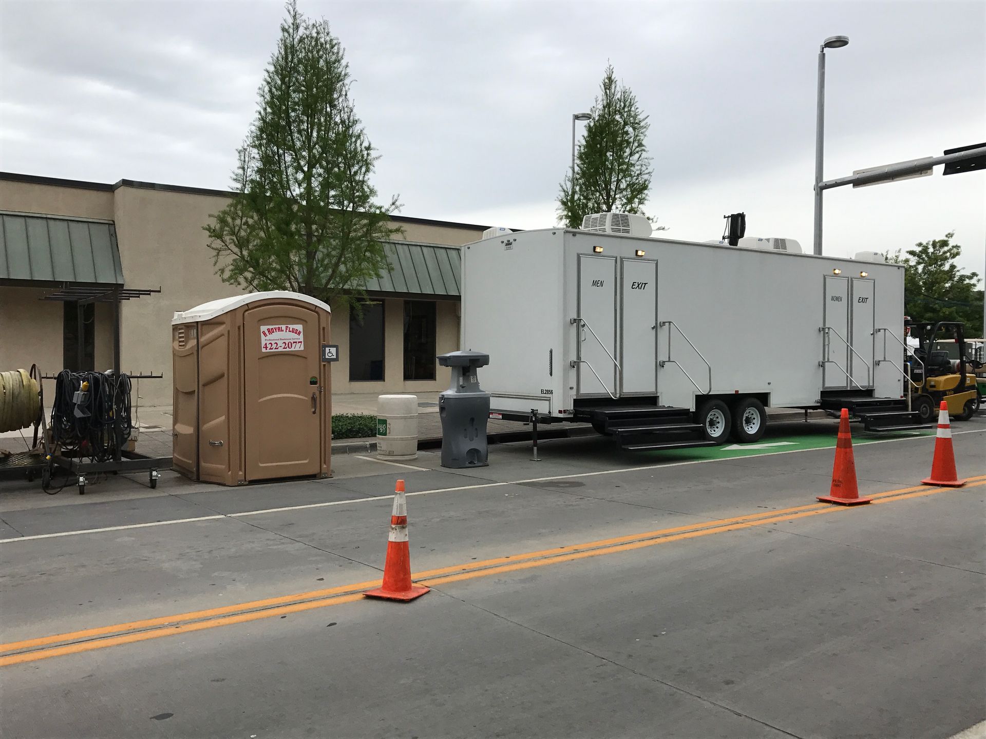 Construction site: portable toilet and trailer beside building on street. Traffic cones delineate work area.