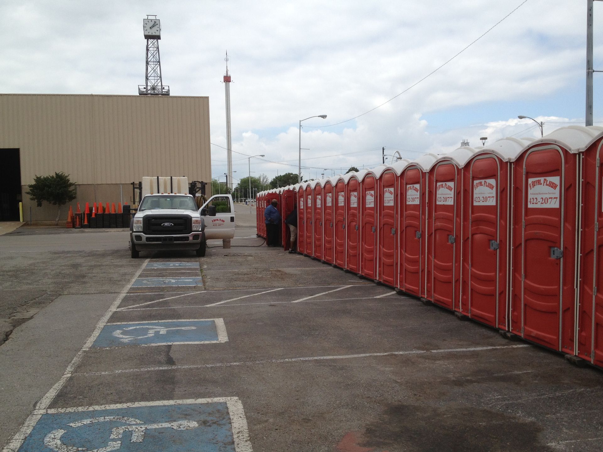 A line of red portable toilets next to a parking area with a truck and building in the background.