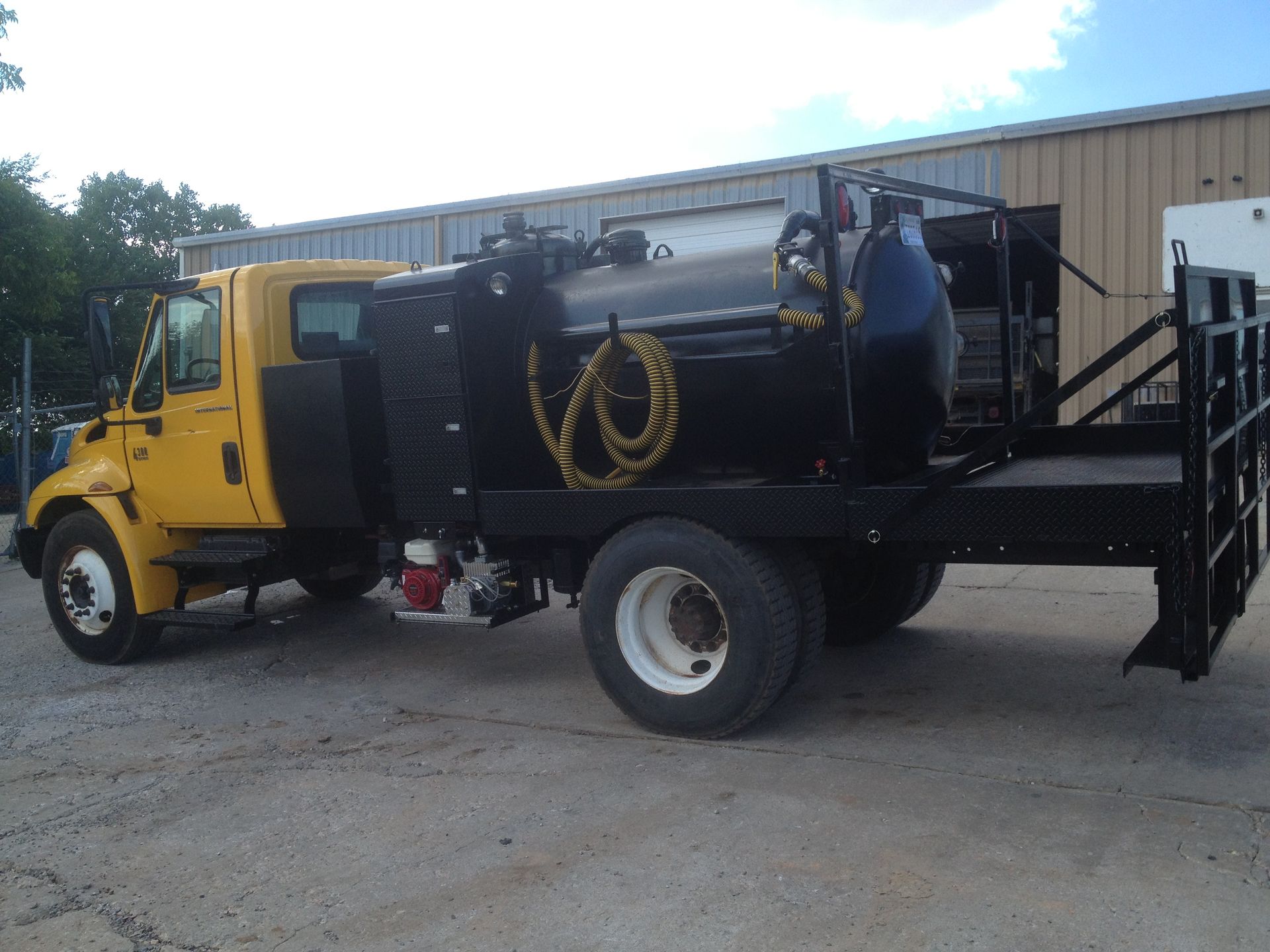 Yellow truck with black tank, hose, and flatbed parked outside a building.