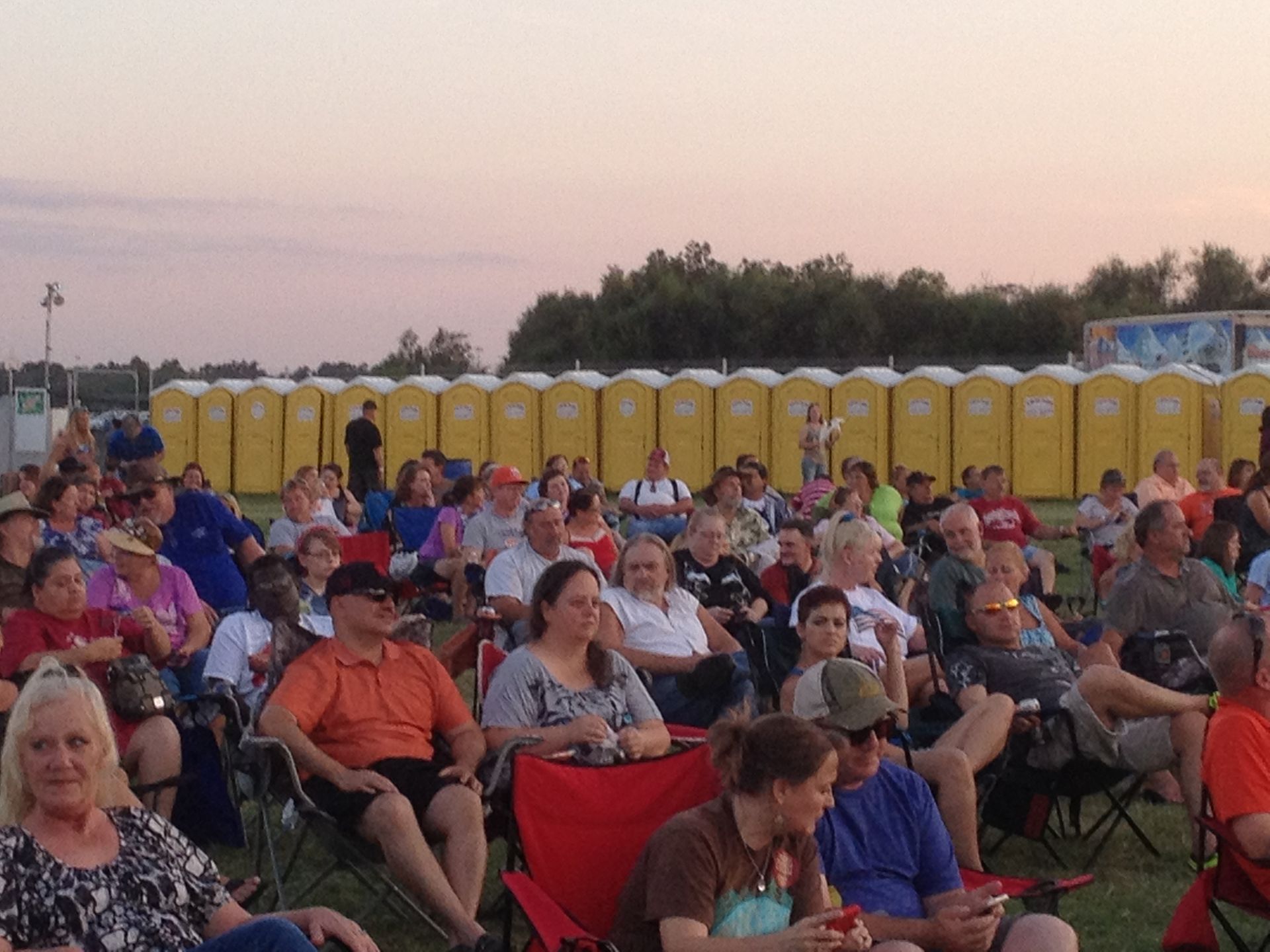 Crowd seated on lawn at an outdoor event, looking toward something out of frame. Yellow portable toilets in the background.