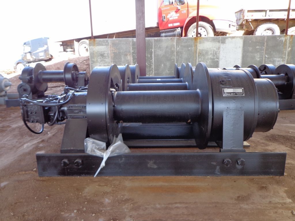A heavy-duty industrial winch assembly in black, sitting on a metal frame at an outdoor job site.