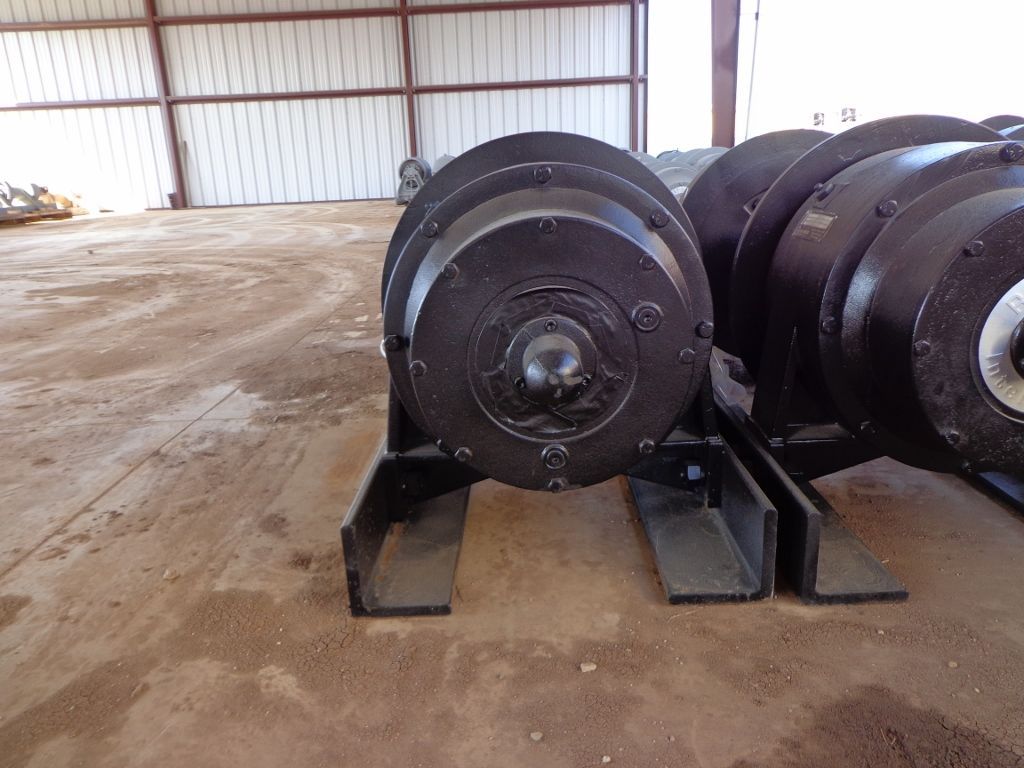 Two black, cylindrical heavy-duty industrial winches mounted on metal frames, situated on a dirt floor inside a hangar.