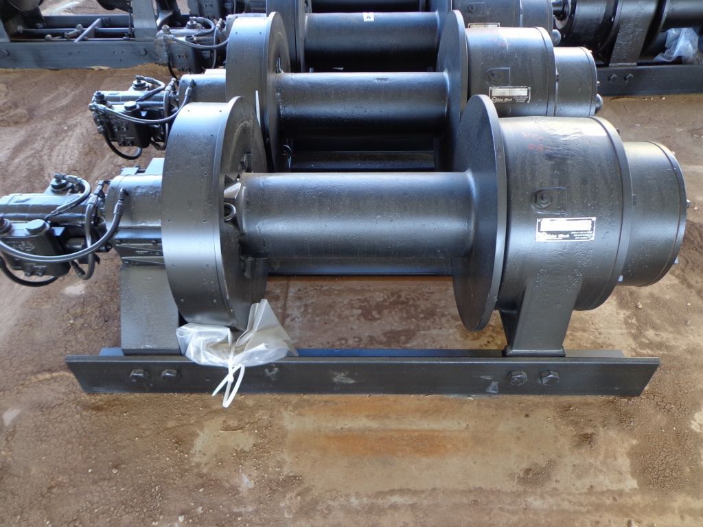 A close-up view of several black industrial hydraulic winches mounted on a steel base, positioned on a dirt ground.