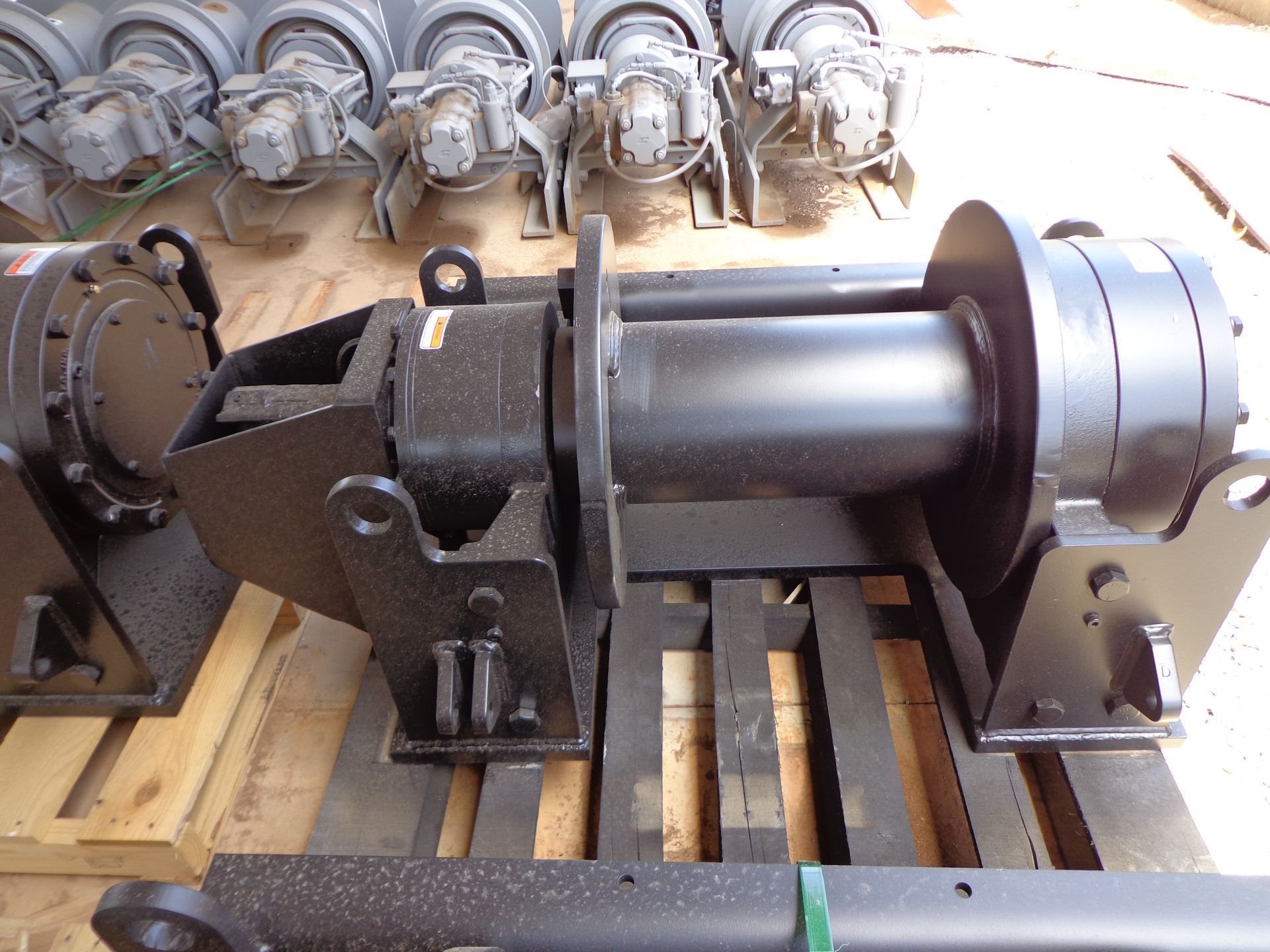 A black industrial hydraulic winch sits on a wooden pallet with several matching units visible in the background.