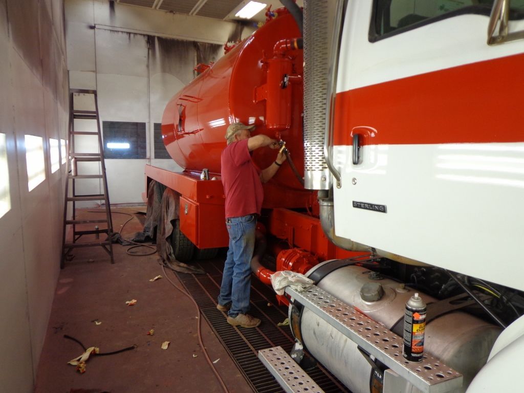 Worker repairing a red tanker truck inside a garage with a ladder nearby